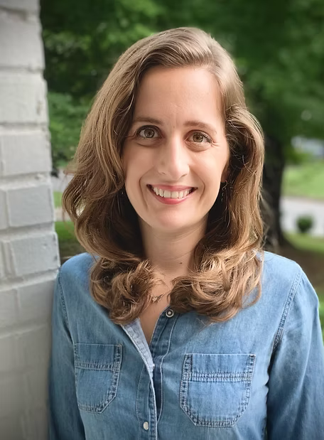 A woman with shoulder-length wavy brown hair and a bright smile, wearing a denim shirt, standing outdoors near a white brick wall with green trees in the background.