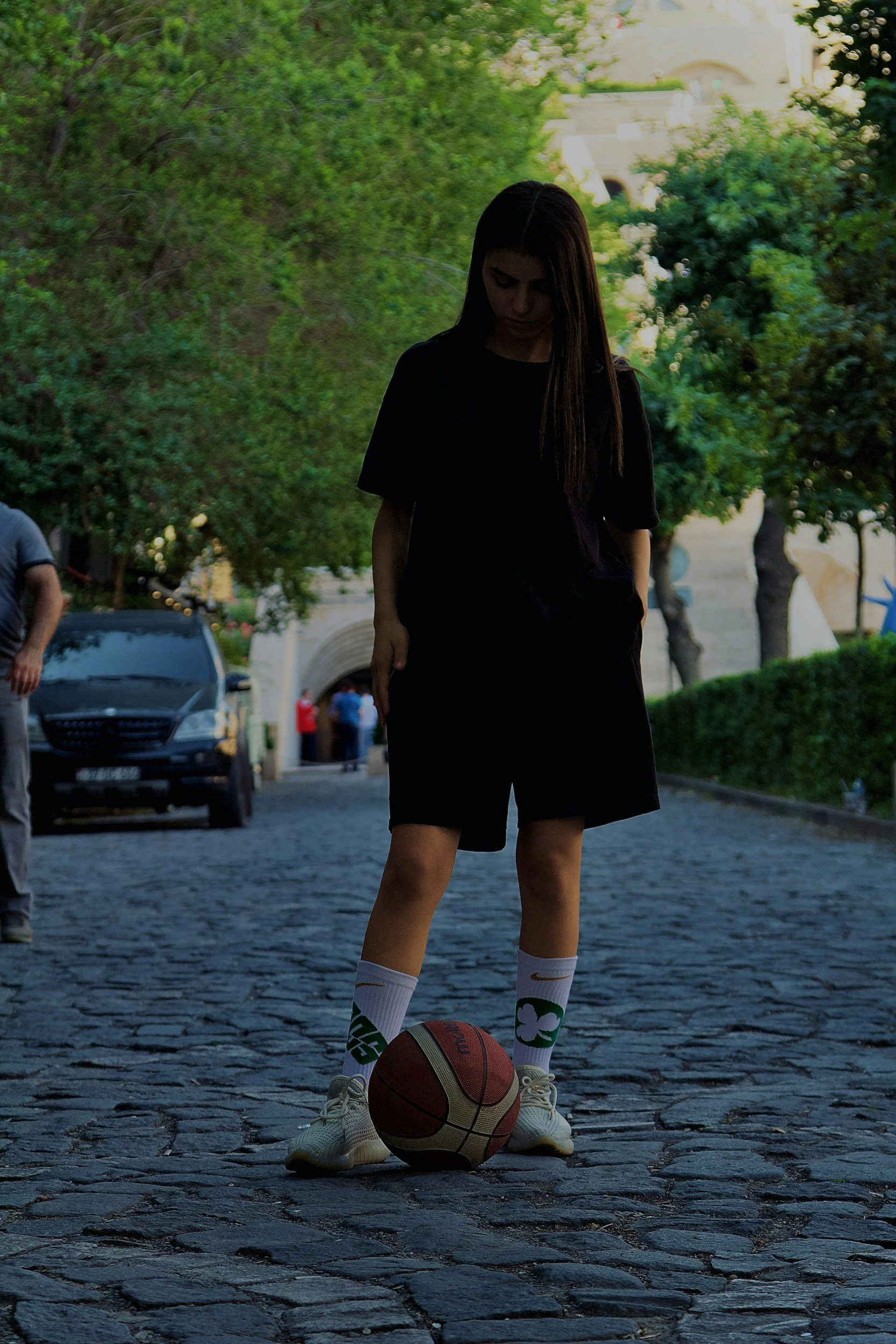 A young woman in black clothing and white Nike socks with shamrock and green logo, standing on cobblestone street with a basketball at her feet, outdoors with trees and buildings in the background.
