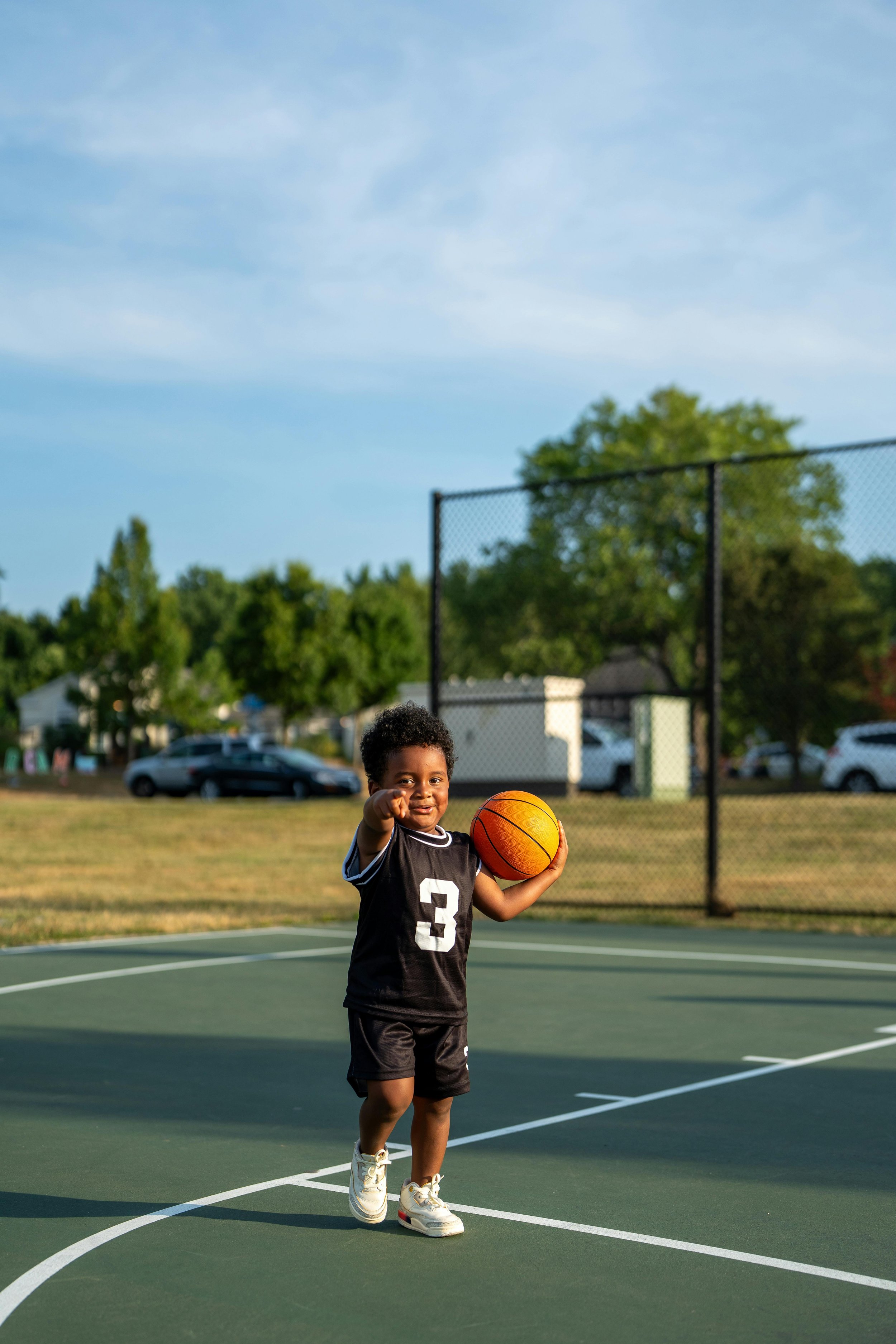 A young child on a basketball court holding a basketball and pointing towards the camera with a smile.
