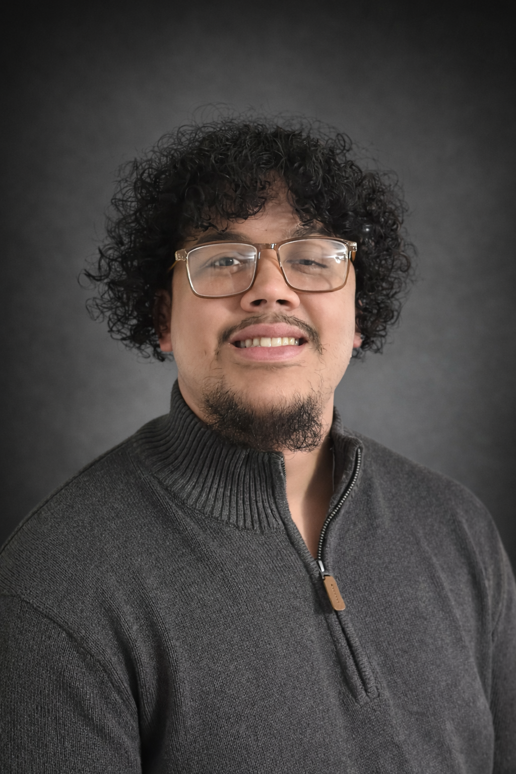 Portrait of a man with curly black hair, wearing glasses, and a dark grey zip-up sweater, standing against a dark background.