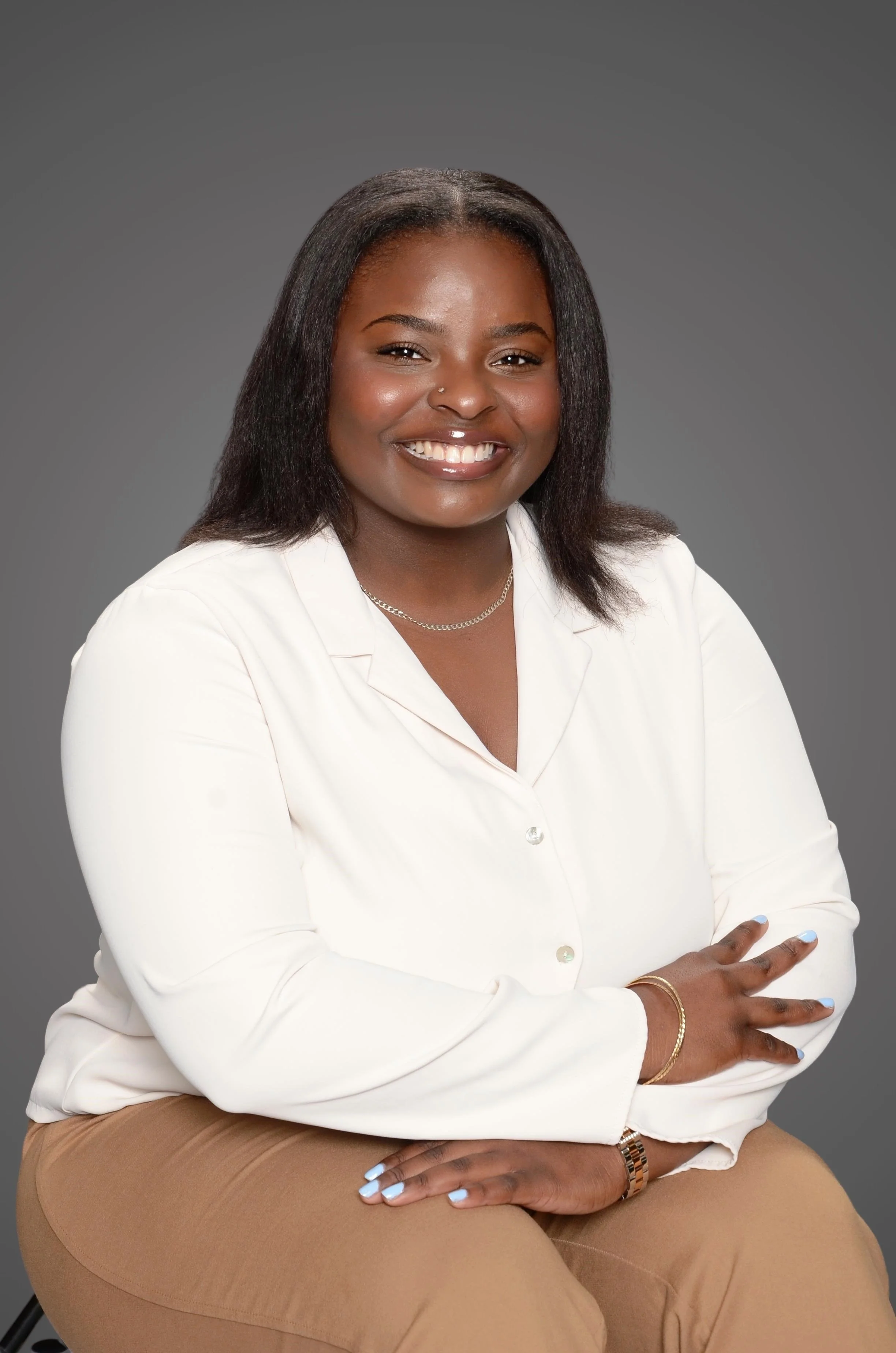 A smiling woman sitting with arms crossed against a gray background, wearing a white blouse and tan pants.