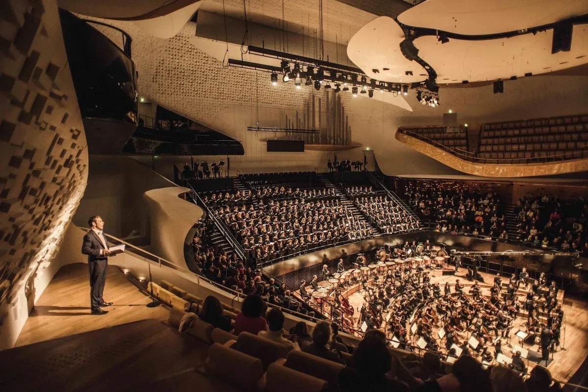 An orchestra performing in a modern concert hall with an audience seated in tiered rows.