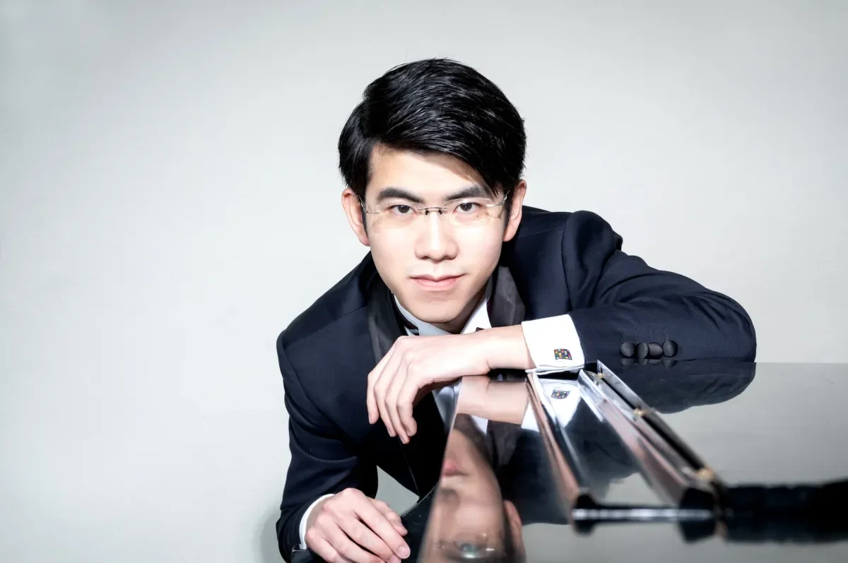 A young man with glasses in a black suit leaning on a piano with a plain light background.