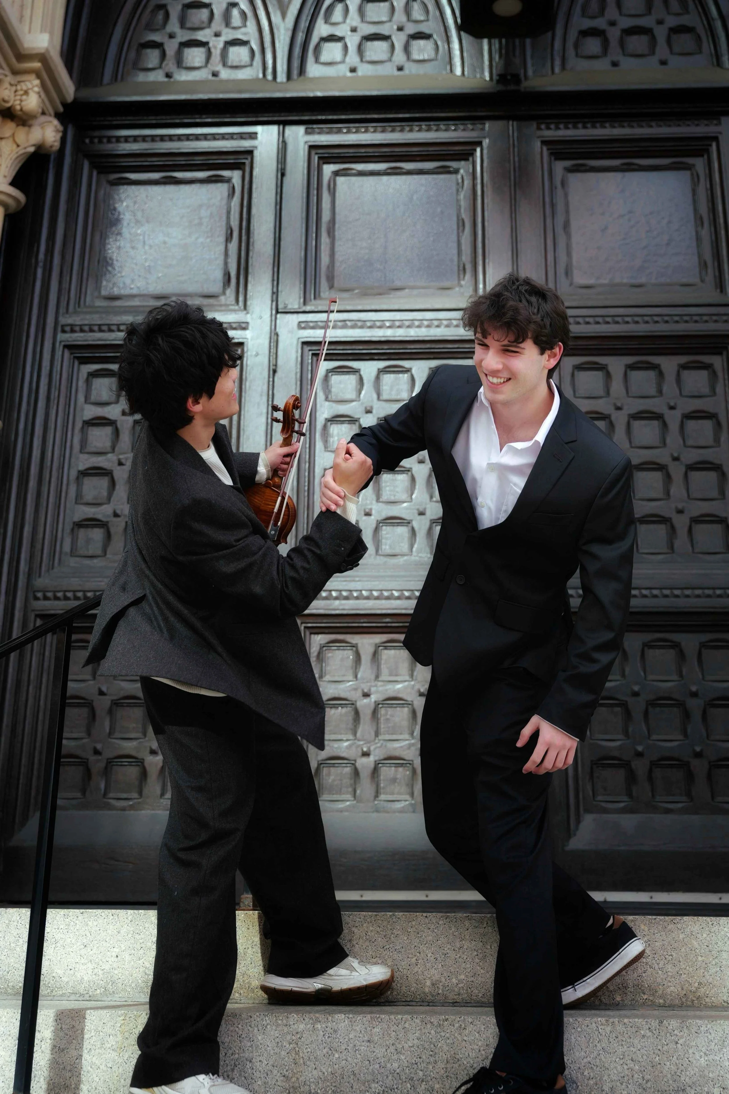Two young men in formal attire smiling and engaging in a playful wrestling match on steps in front of a ornate wooden door.