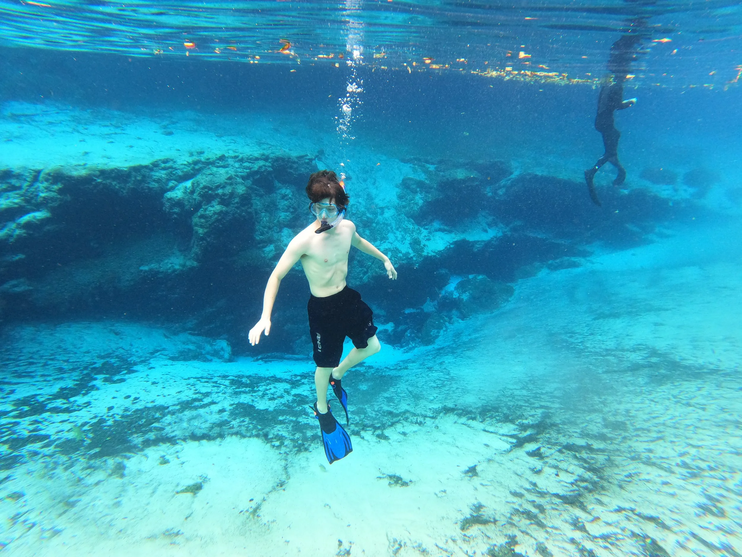 An underwater snapshot of a young man snorkeling deep below the surface, he is surrounded by crystal clear fresh water, behind him an underwater cave is present and another diver's legs can be seen almost out of frame.