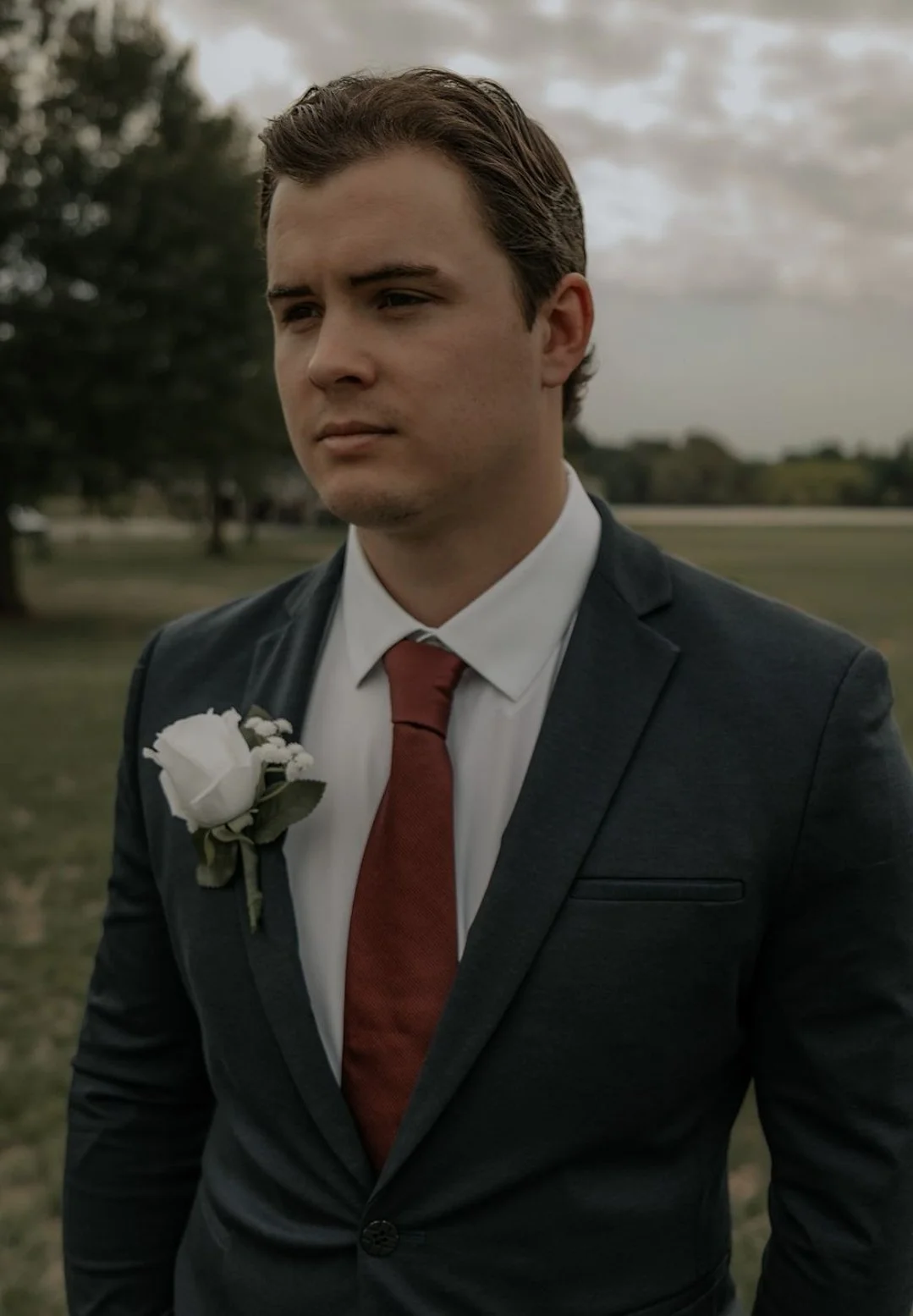 A young man in a dark suit, white shirt, and red tie standing outdoors with trees and cloudy sky in the background, wearing a white flower boutonniere on his lapel.
