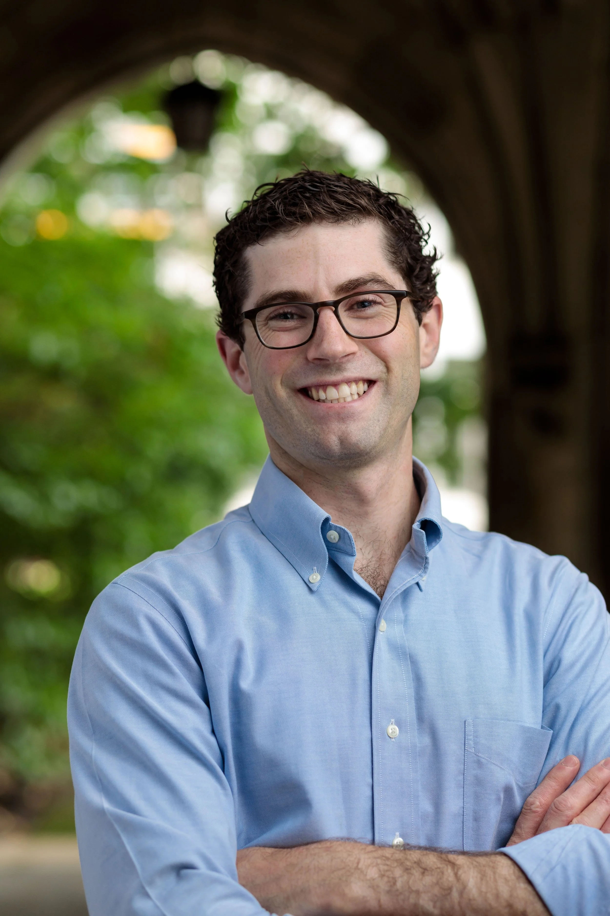 A smiling man with glasses and dark curly hair, wearing a light blue button-up shirt, standing outdoors with green trees and an archway in the background.
