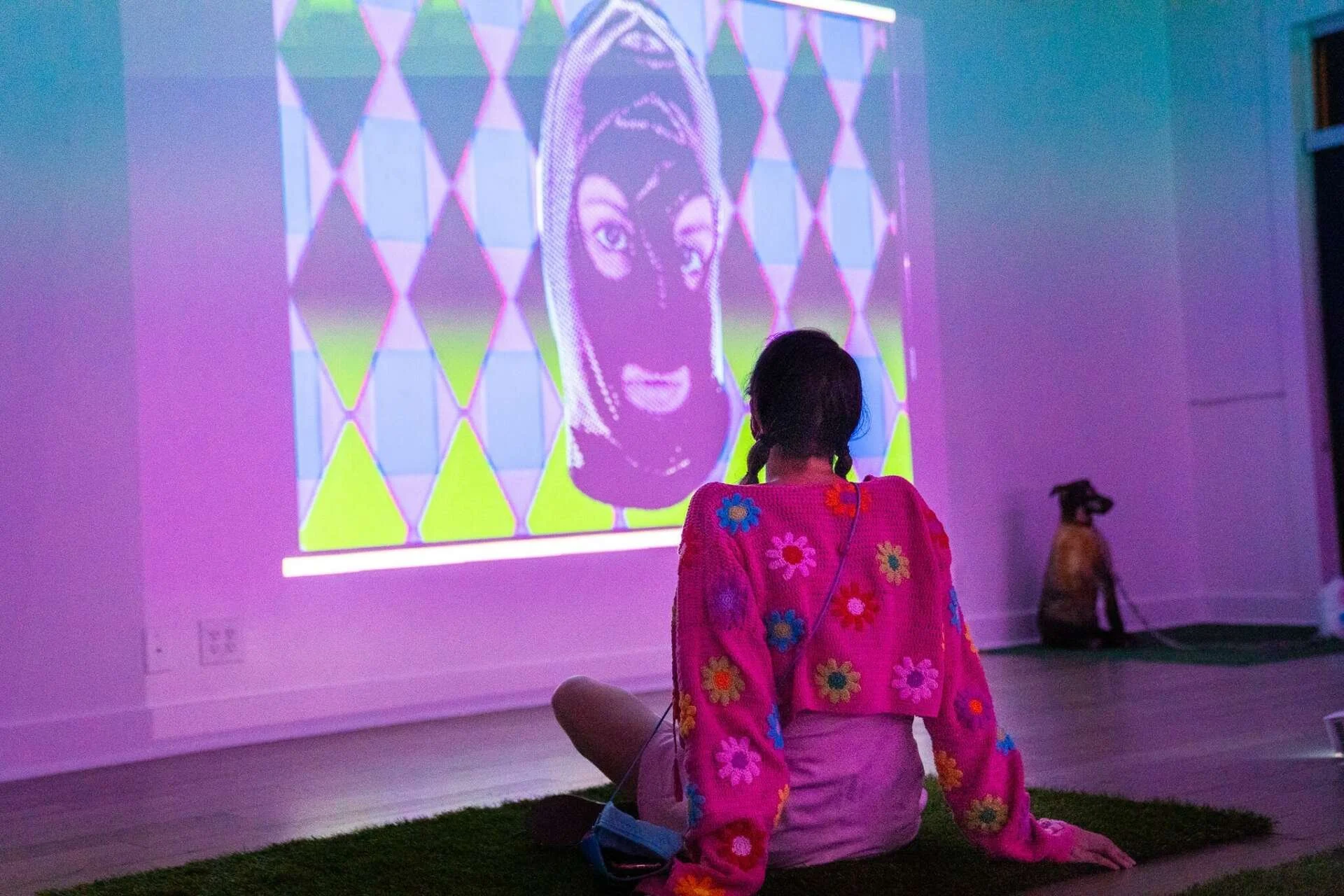 A girl with braided hair wearing a colorful floral sweater sits on the floor watching a digital art projection on the wall, which depicts a stylized face with a pattern of geometric shapes in purple, green, and blue tones. There is a dog sitting near