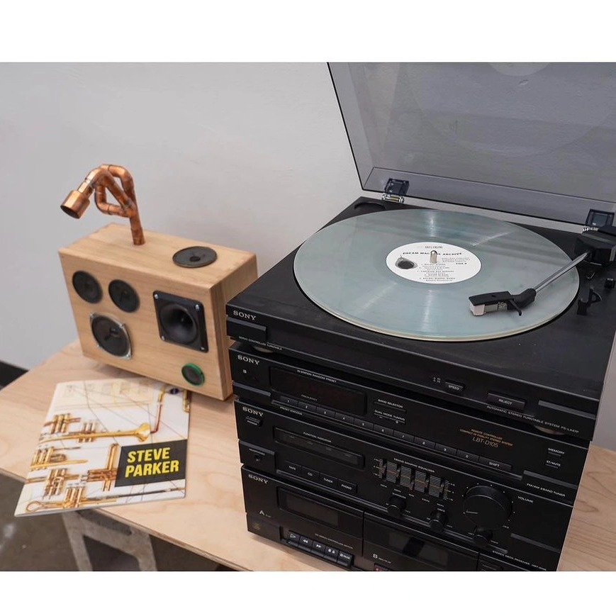 Turntable with vinyl record playing, a stack of Sony audio equipment, a wooden speaker with copper piping, and a book titled End of the World by Steve Parker on a wooden table.