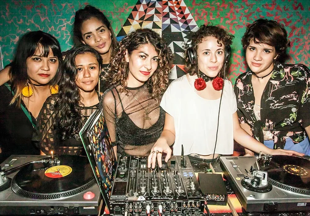 Group of seven women DJing and posing behind turntables in a colorful, patterned room.