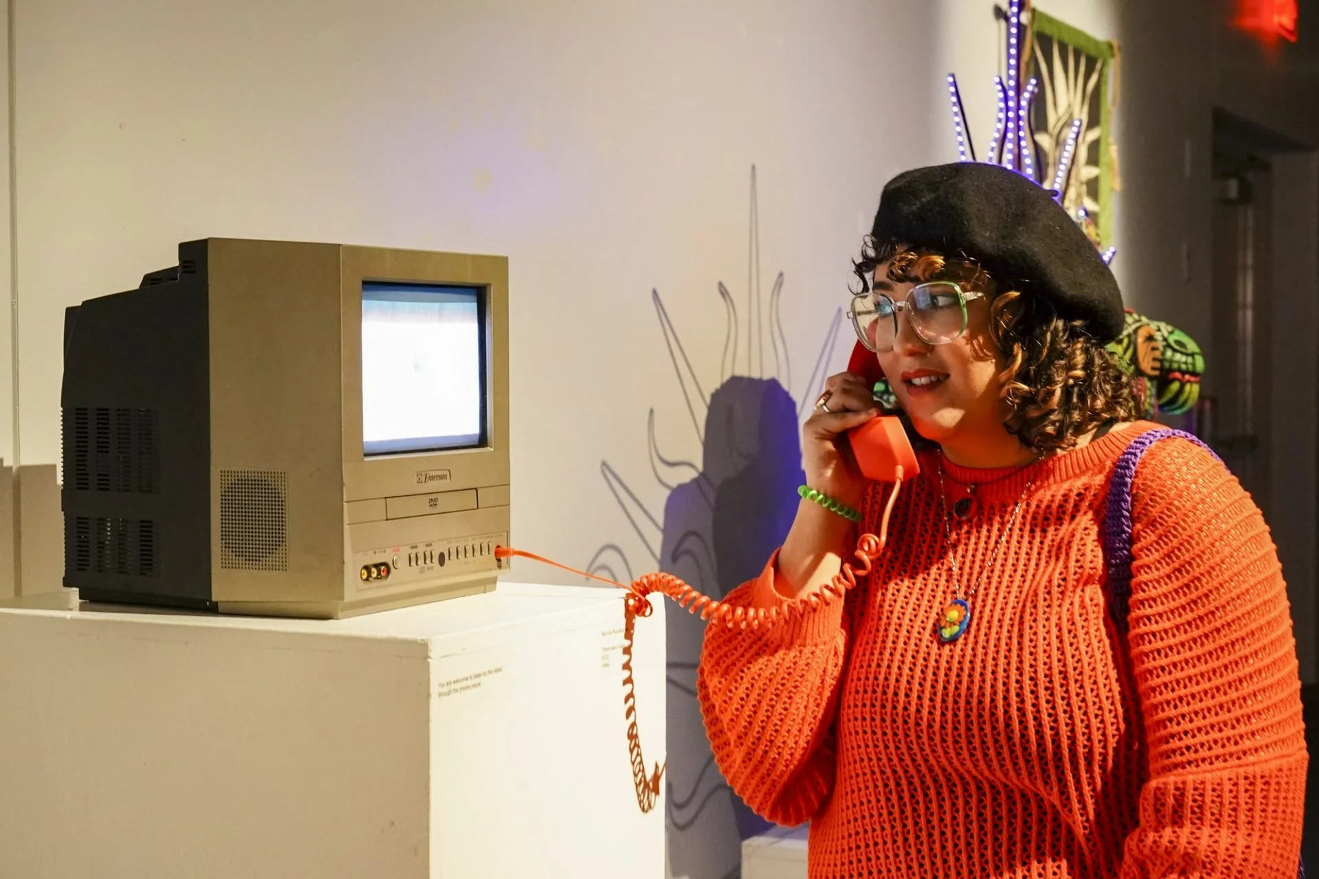 Young woman with glasses, curly hair, black beret, orange sweater, and colorful jewelry, talking on a red rotary phone connected to an old CRT television, in a vibrant art gallery with colorful décor.