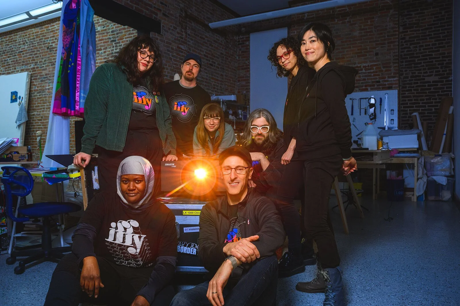 A diverse group of nine people posing together in a workshop or studio with brick walls, some wearing black shirts with the word 'jiv' on them, with a bright light providing a lens flare effect.