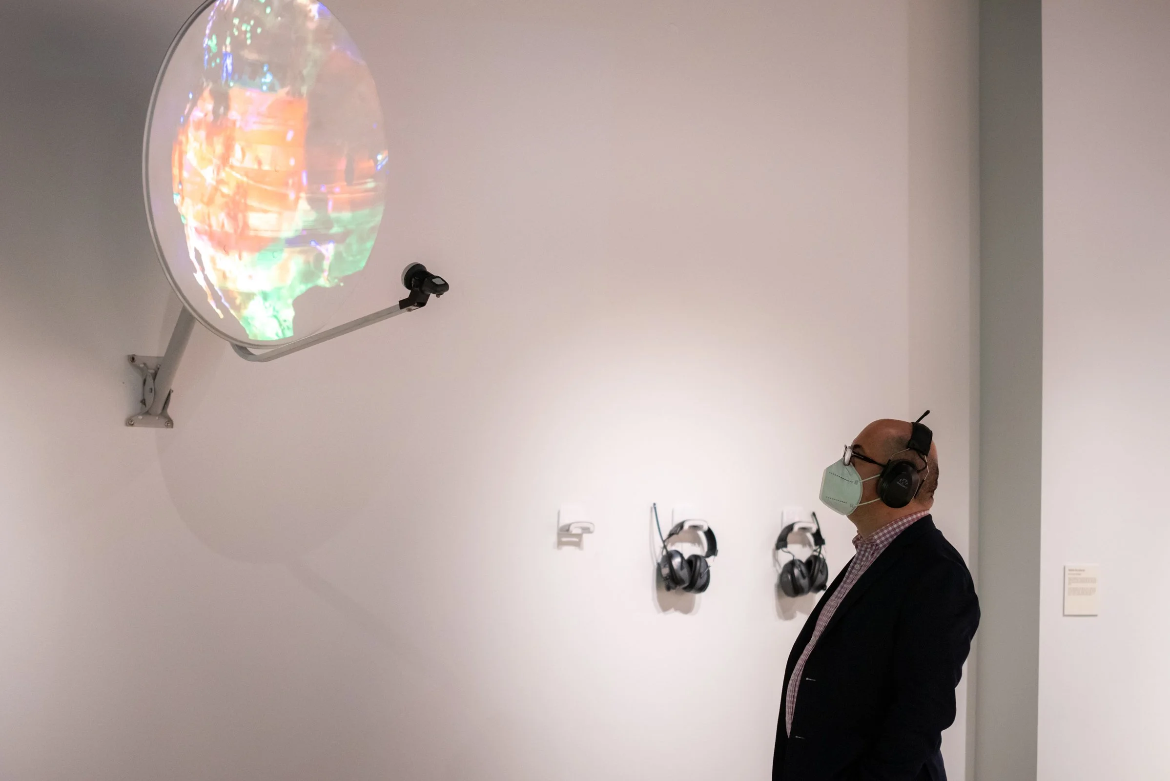 A man wearing a face mask and headphones observing a digital globe display in an art gallery or museum.