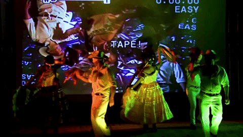 People dancing in front of a large screen displaying the word 'TAPE!' and camera settings.