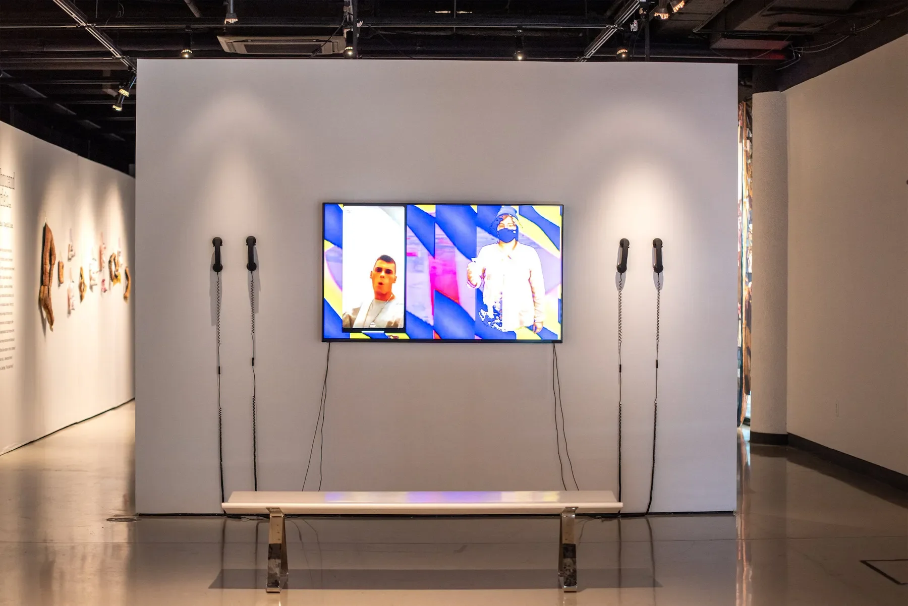 Art exhibit with a mounted TV screen displaying digital artwork, four headphones on either side, and a white bench in front.