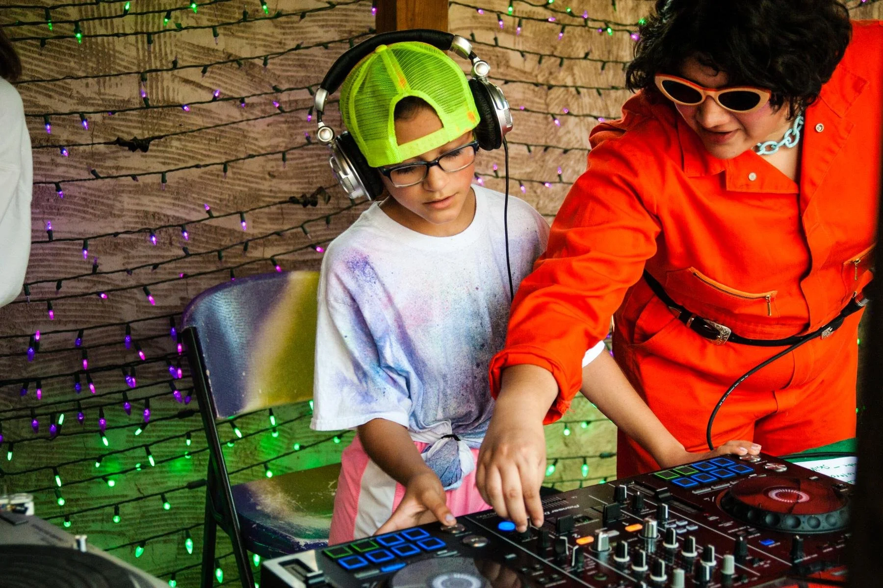 A young person wearing headphones, glasses, and a neon green cap, is standing next to a DJ, operating DJ equipment. The DJ is dressed in an orange jumpsuit and is wearing sunglasses and a chunky necklace. The background has a wooden wall with colorfu
