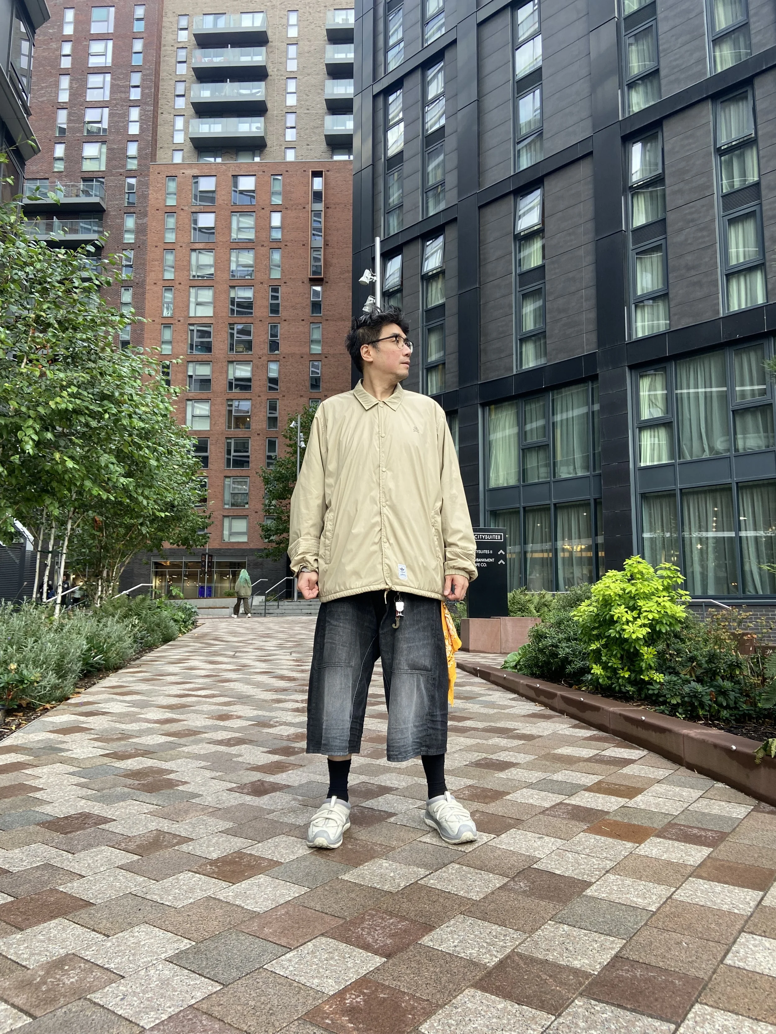 A young man standing on a brick-paved path in an urban area, wearing a beige jacket, dark cropped jeans, black socks, and white sneakers, with modern buildings and greenery in the background.