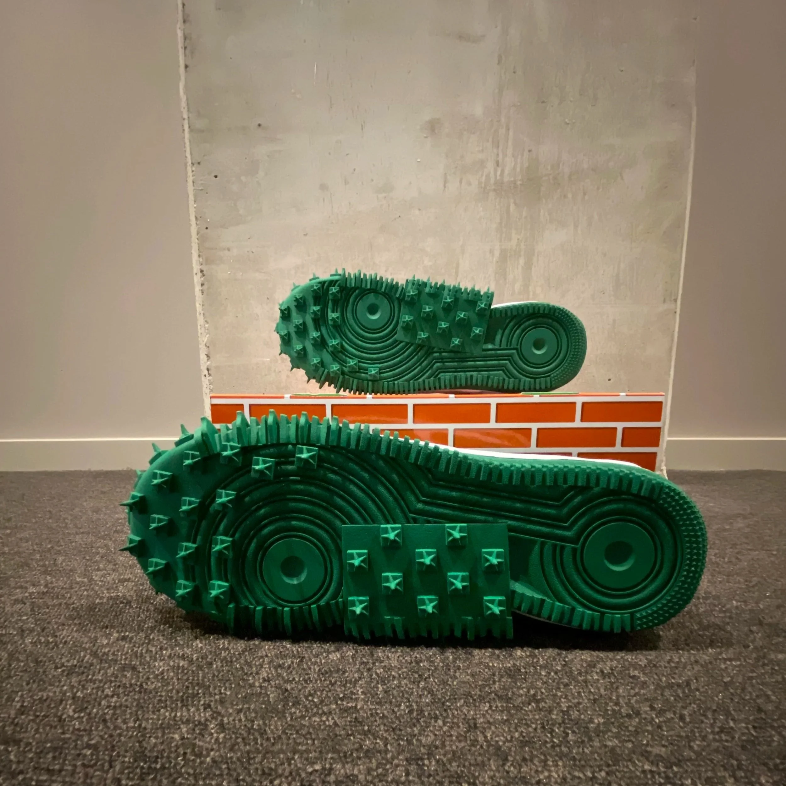 Green rubber shoe soles with spikes and grooves, placed on a dark carpeted floor in front of a wall with a brick pattern and a mirror.