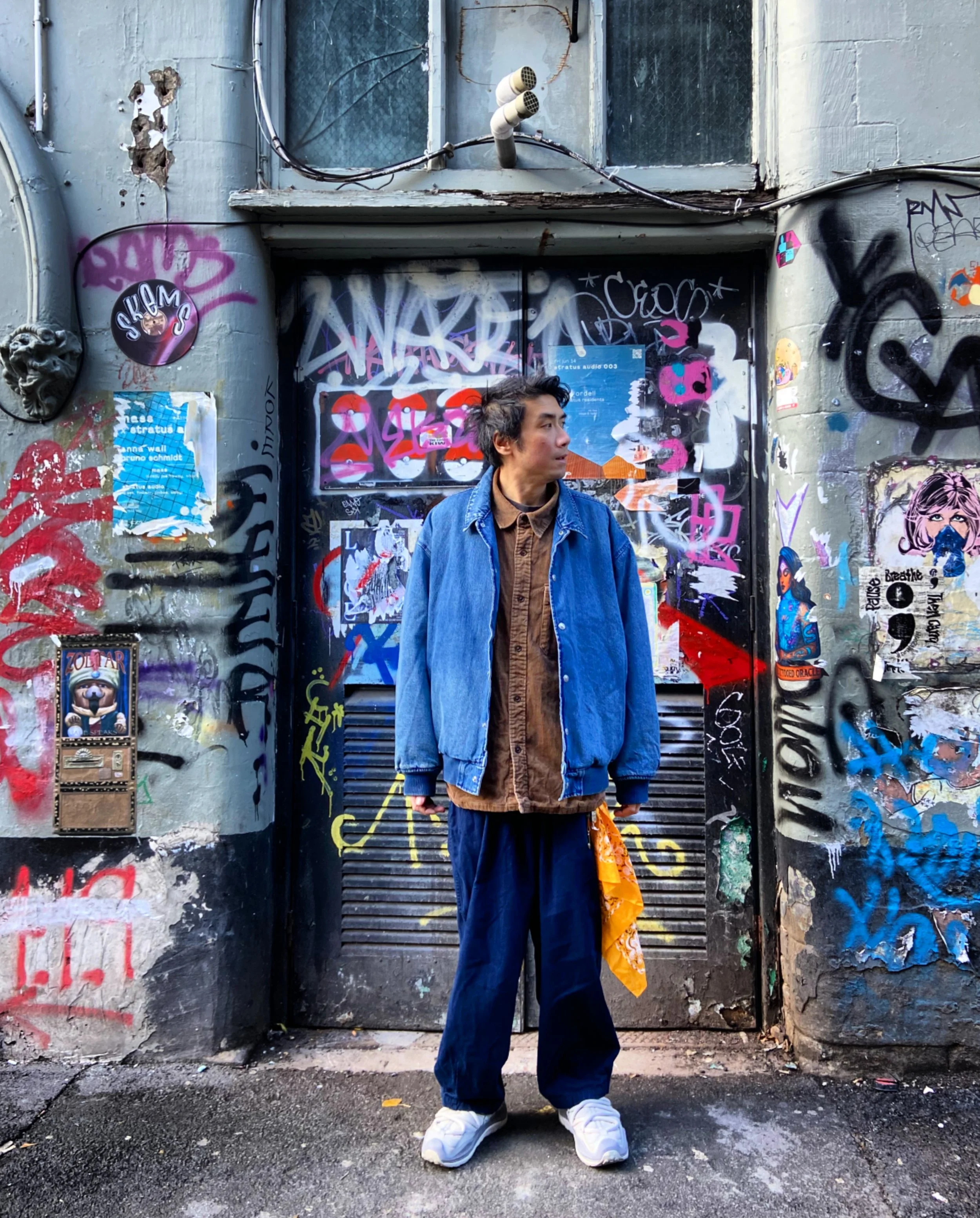 A man in a denim jacket and brown shirt standing against a graffiti-covered wall in an urban setting.