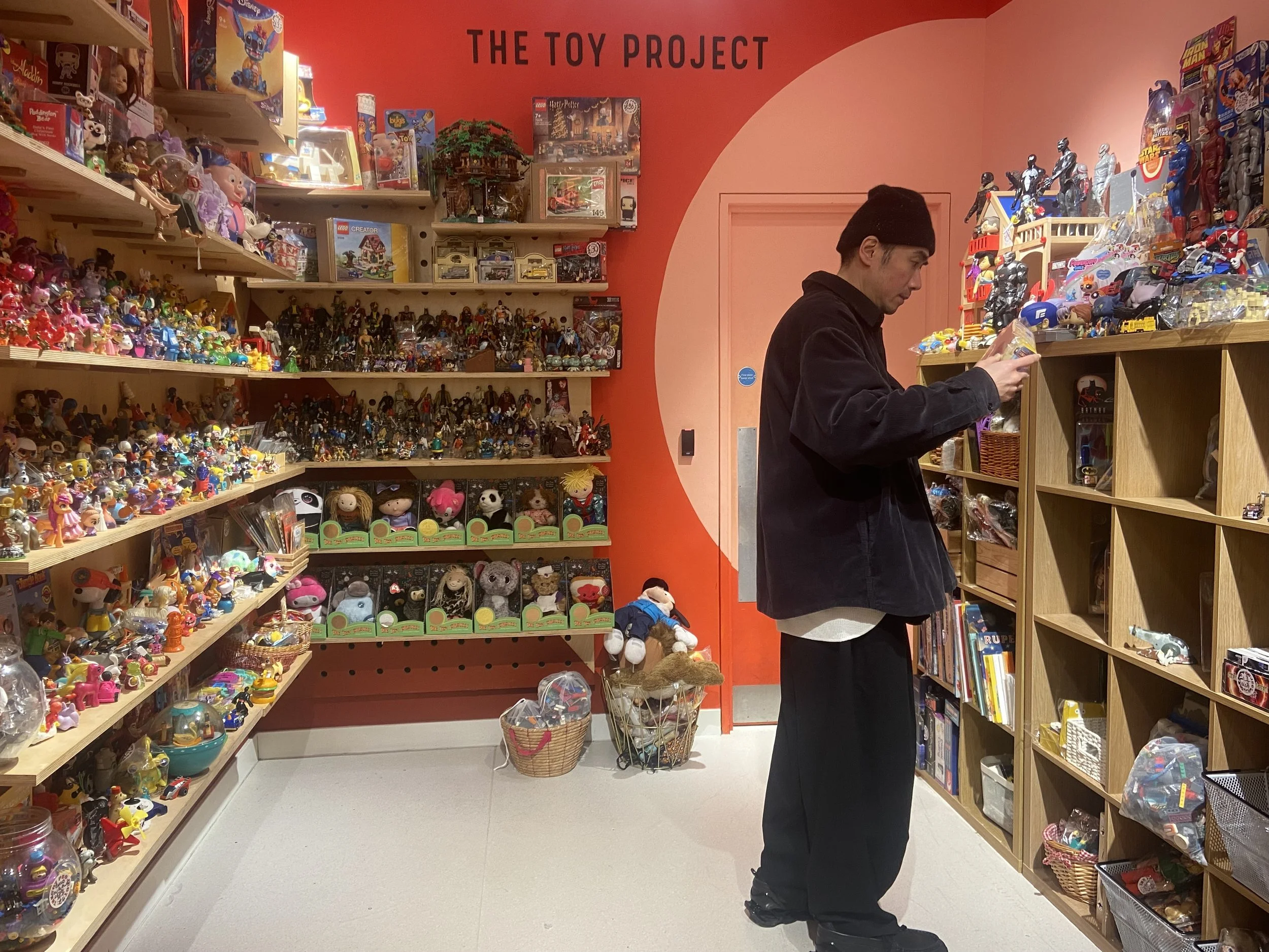 A man in a black jacket and black beanie shopping for toys at a store called 'The Toy Project'. The store has shelves filled with various toys and collectibles, including figurines, plush toys, and action figures.