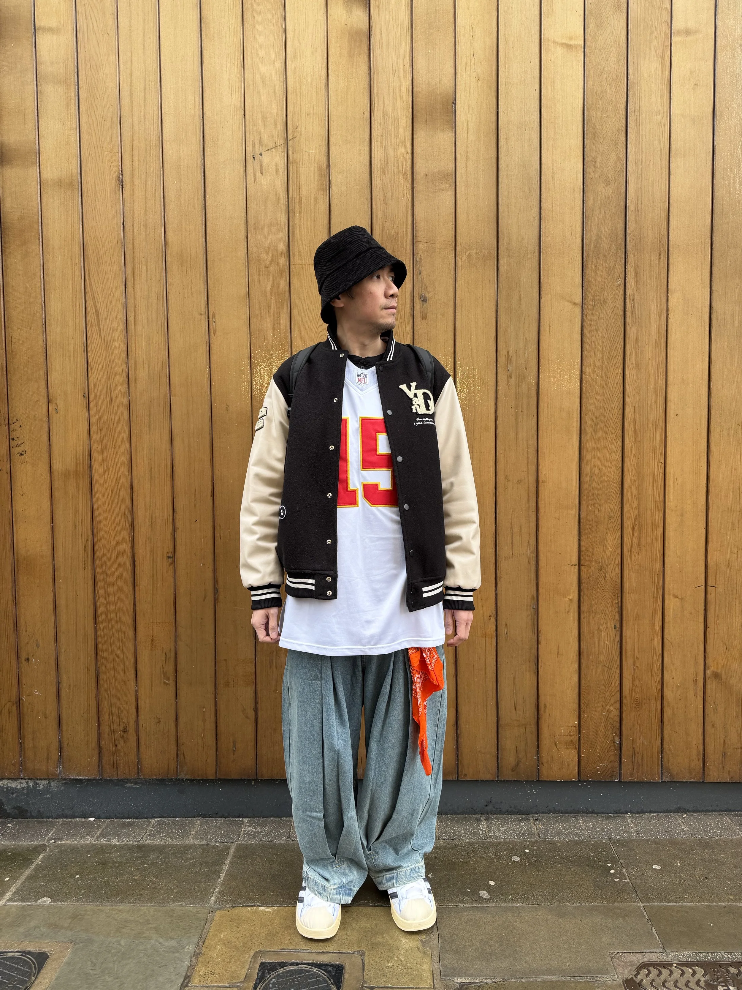 A man stands against a wooden wall, wearing a black and beige varsity jacket, a white football jersey with the number 15, loose blue jeans with a red bandana tied to the pocket, white sneakers, and a black bucket hat.