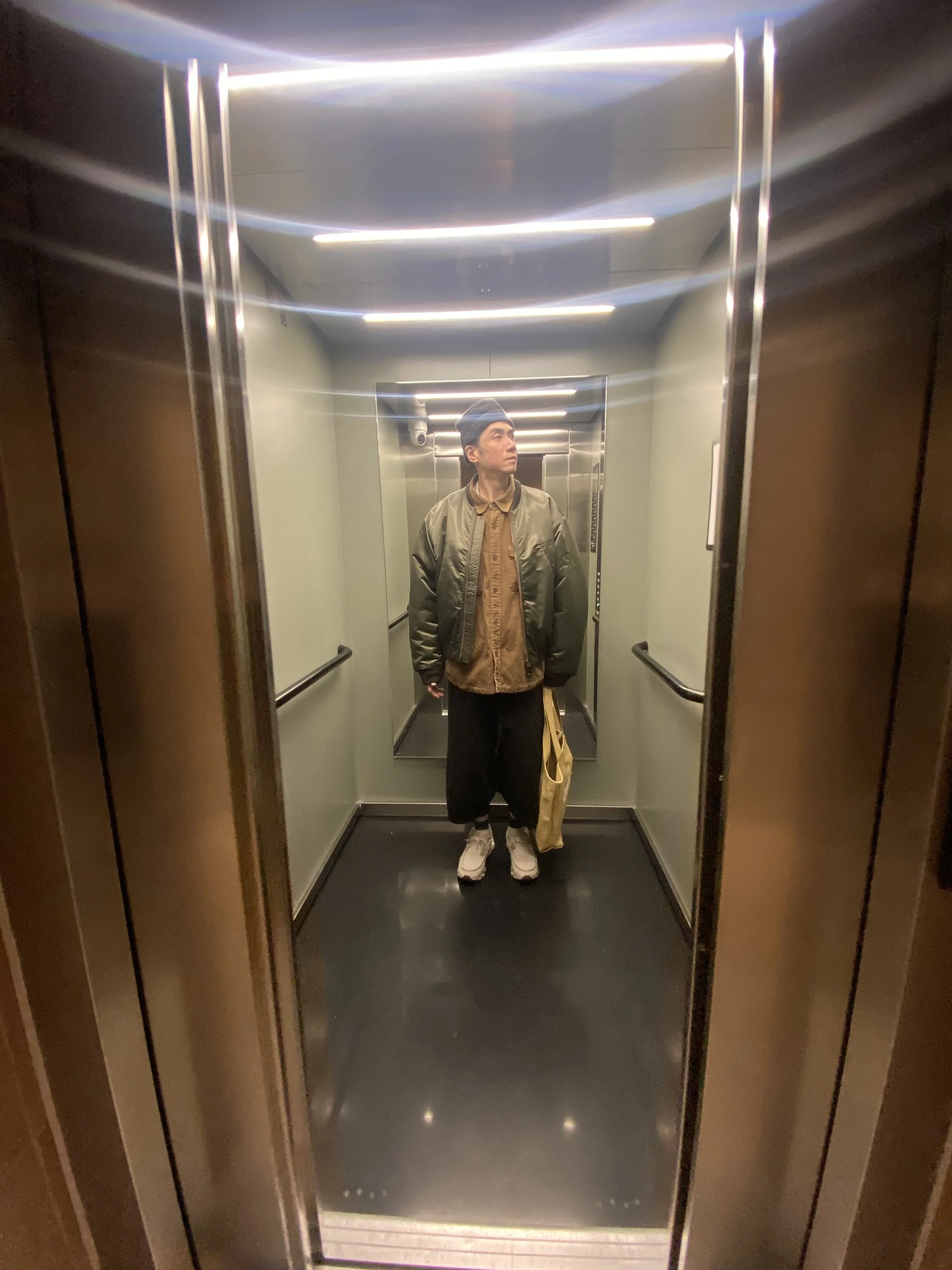 Person standing in an elevator, looking to the side, holding a beige bag, wearing a green bomber jacket, beige shirt, black pants, white sneakers, and a gray beanie.