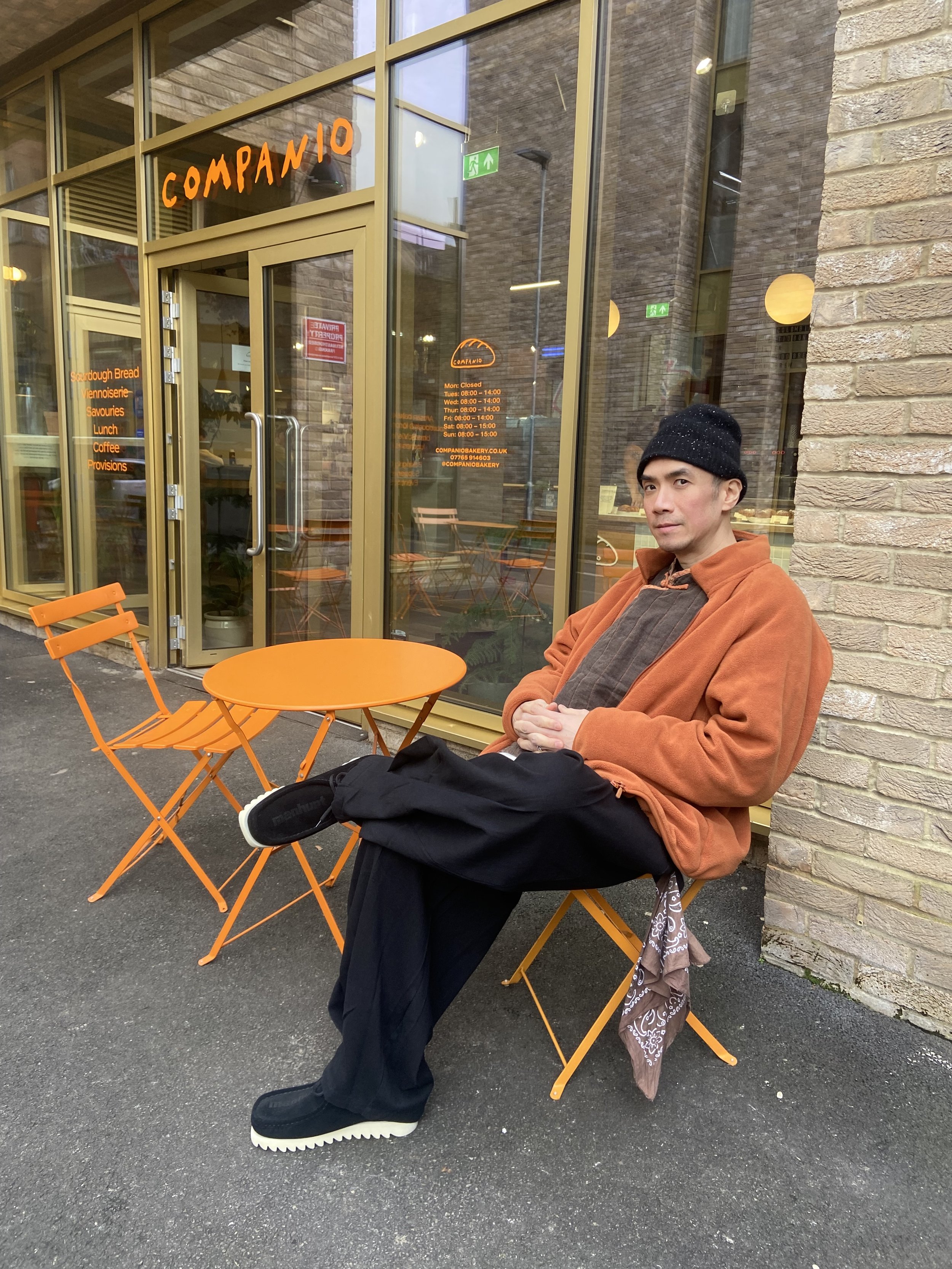 A man sitting outside a bakery with a brick and glass storefront, orange chairs, and a round table. He is wearing a black beanie, orange jacket, dark pants, and black shoes, looking at the camera.