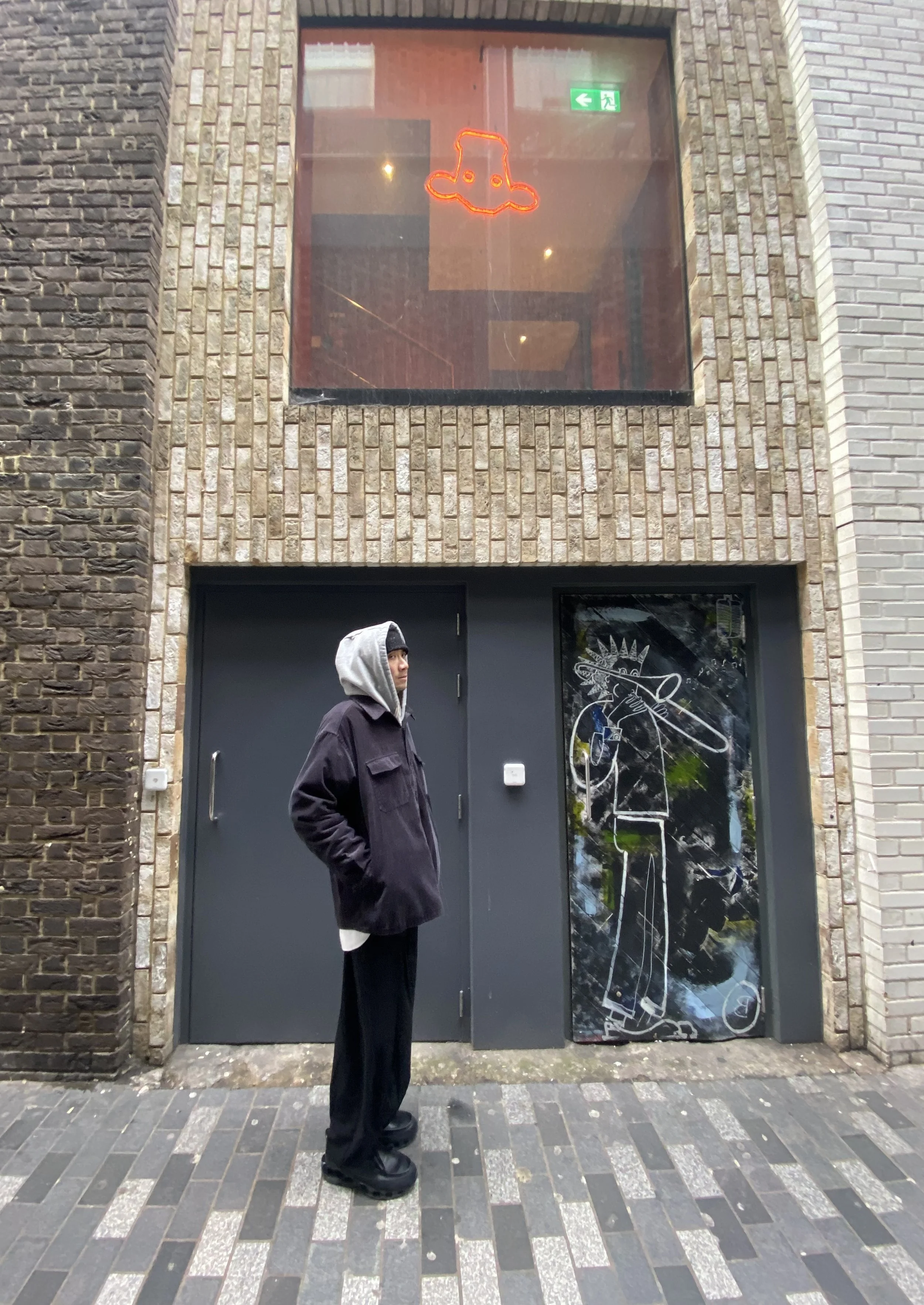 A person dressed in dark clothing with a grey hoodie standing on a sidewalk outside a brick building. The building has a black door with a glass panel featuring a white outline drawing of a cowboy with a hat, pointing a gun. Above, there is a glass window with a neon red and orange outline of a face wearing a hat, along with a green emergency exit sign.