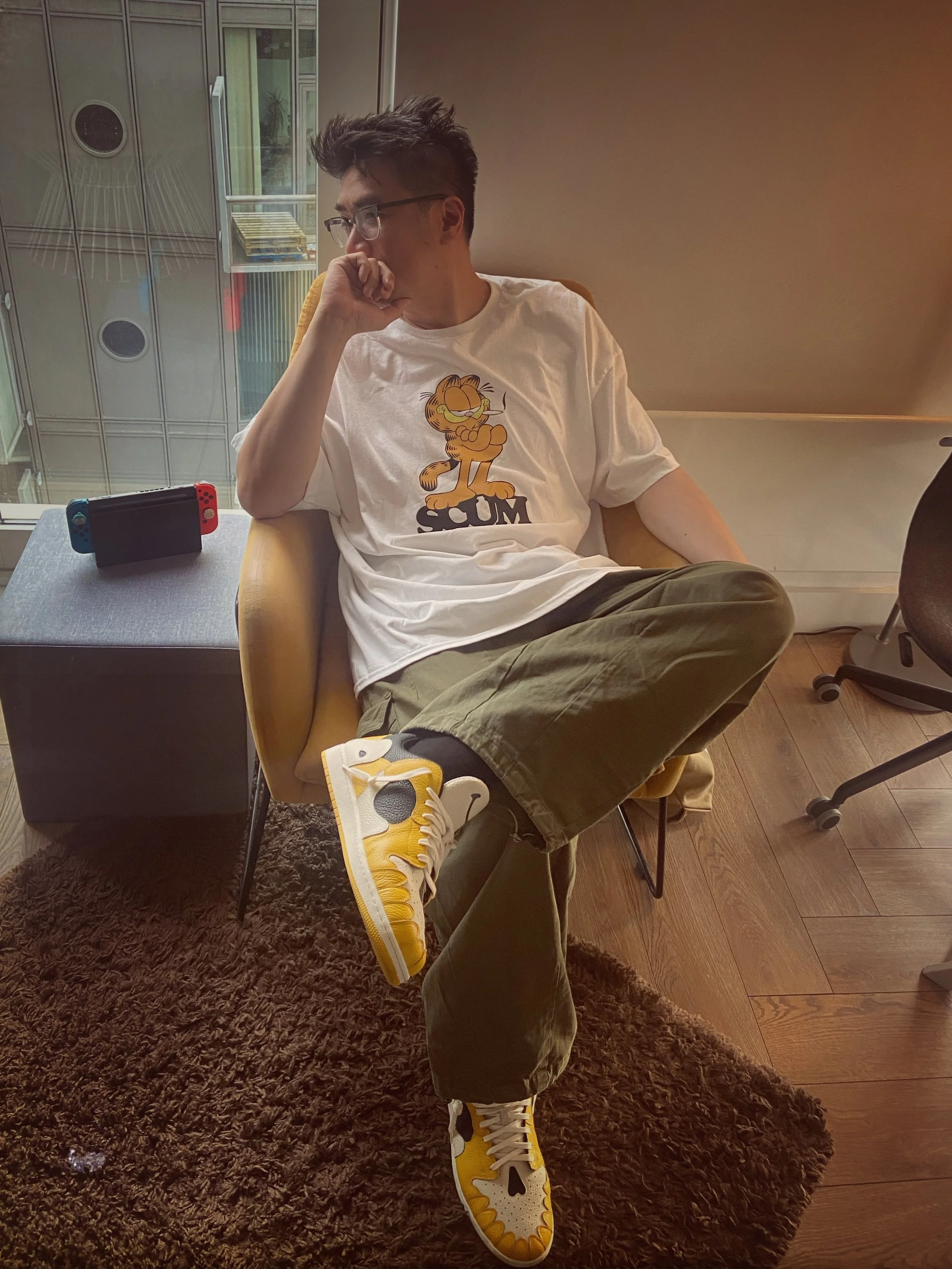 A young man with glasses and styled hair sitting in a yellow armchair, wearing a white Garfield t-shirt, khaki pants, and yellow and white sneakers, with his right hand covering his mouth and his left leg crossed over his right knee. There is a Nintendo Switch on a side table next to him and a window with a balcony in the background.