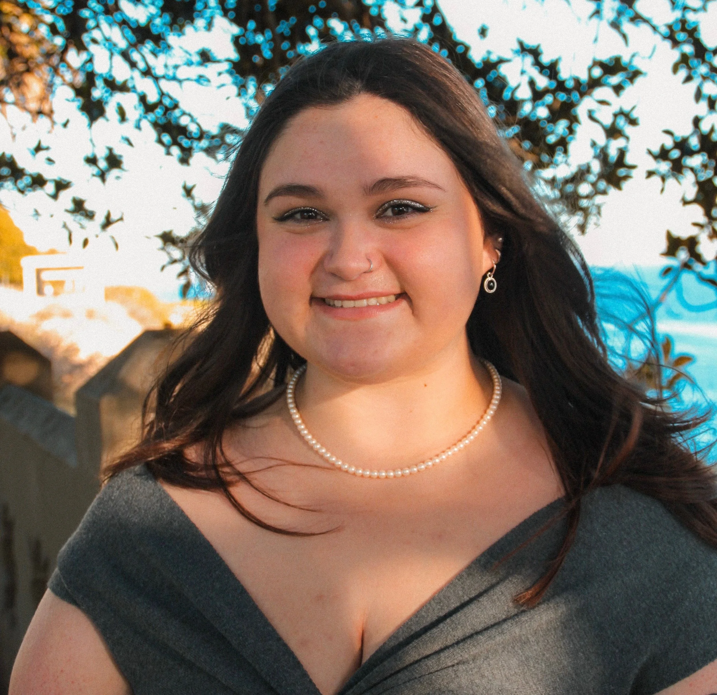 A young woman with dark hair smiling outdoors, wearing pearl jewelry and a grey dress, with trees and a body of water in the background.
