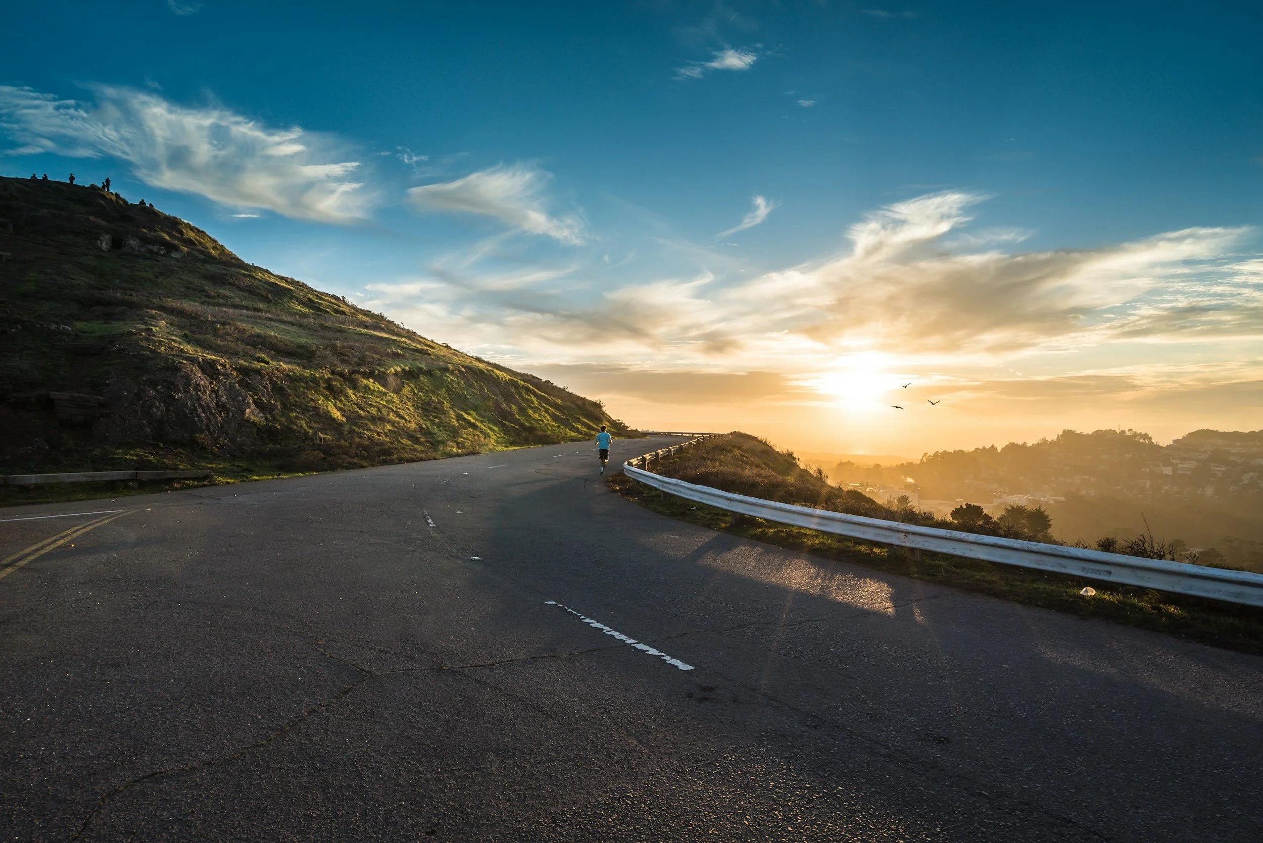 Person jogging on a mountain road at sunrise, with a blue sky, scattered clouds, and a scenic valley in the background.