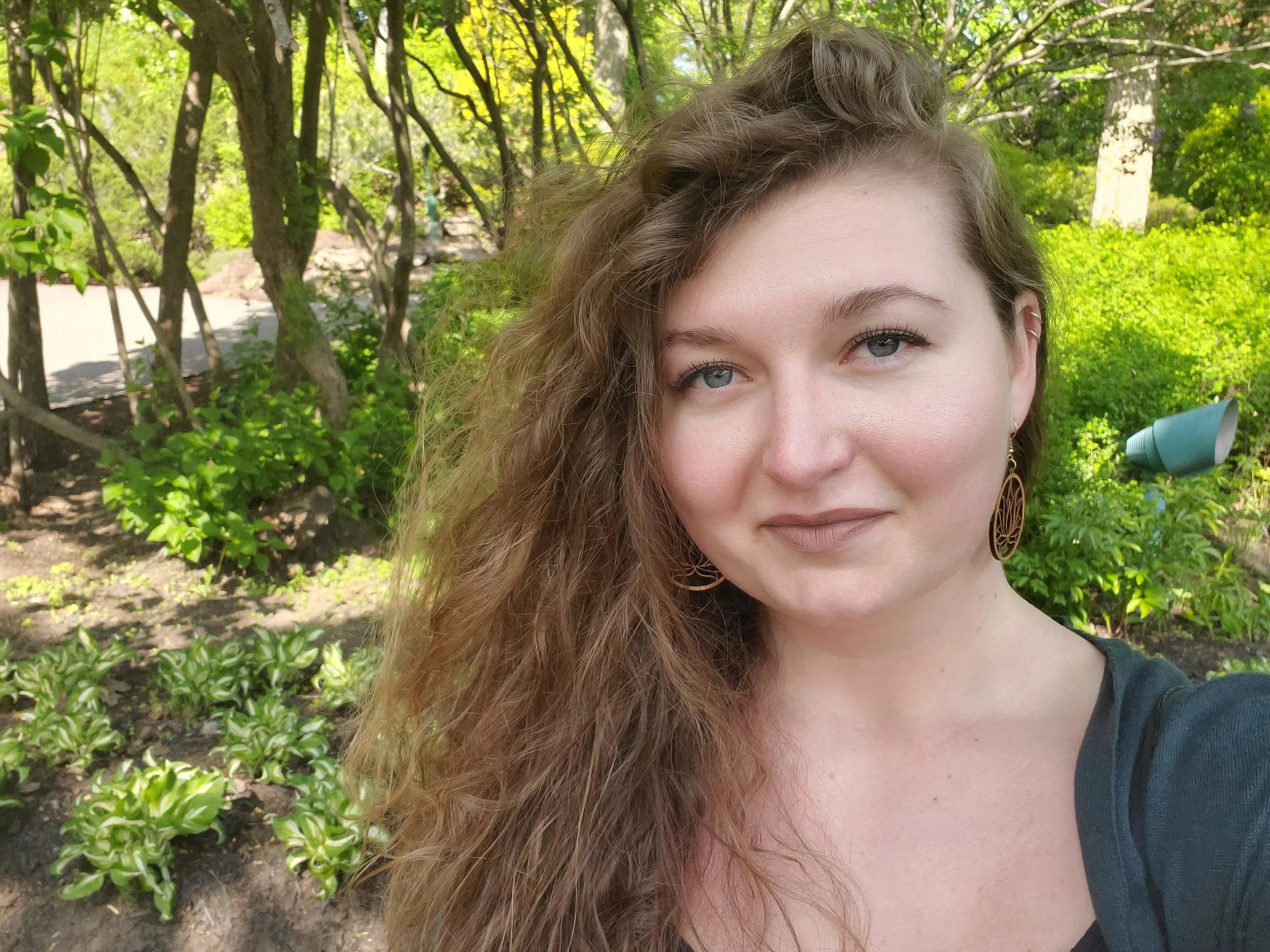 A woman with long, curly hair and blue eyes taking a selfie outdoors, with green trees and plants in the background.