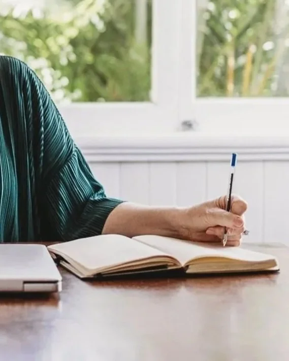 Person sitting at a desk writing in an open notebook with a pen, next to a closed laptop, with a bright window and green foliage outside in the background.