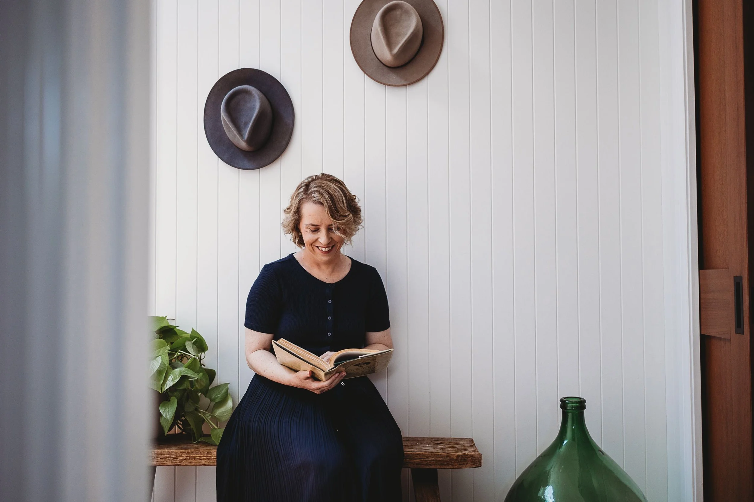 A woman with short blonde hair wearing a black dress is sitting on a wooden bench, reading a book. Behind her, there are two hats hanging on a white paneled wall and a green leafy plant on her left. A large green glass bottle is on the floor beside her.