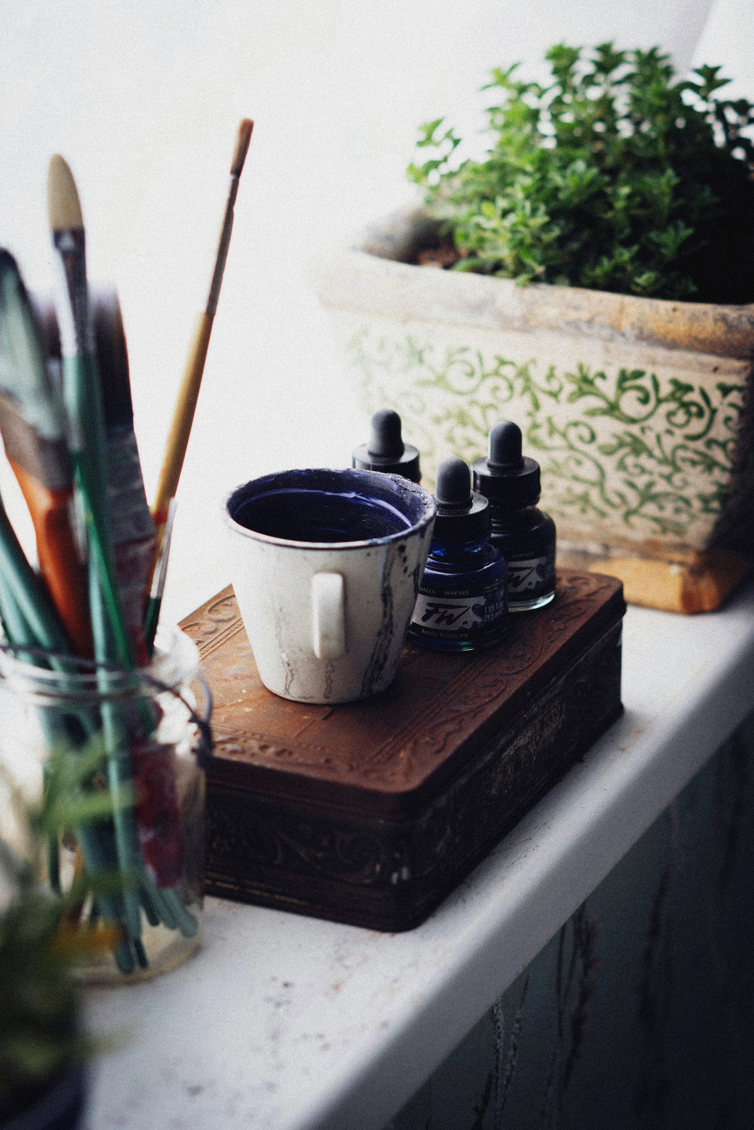 A workspace with paint brushes, a mug with purple paint, three bottles of blue ink, a potted plant, and a decorative wooden box on a white surface.
