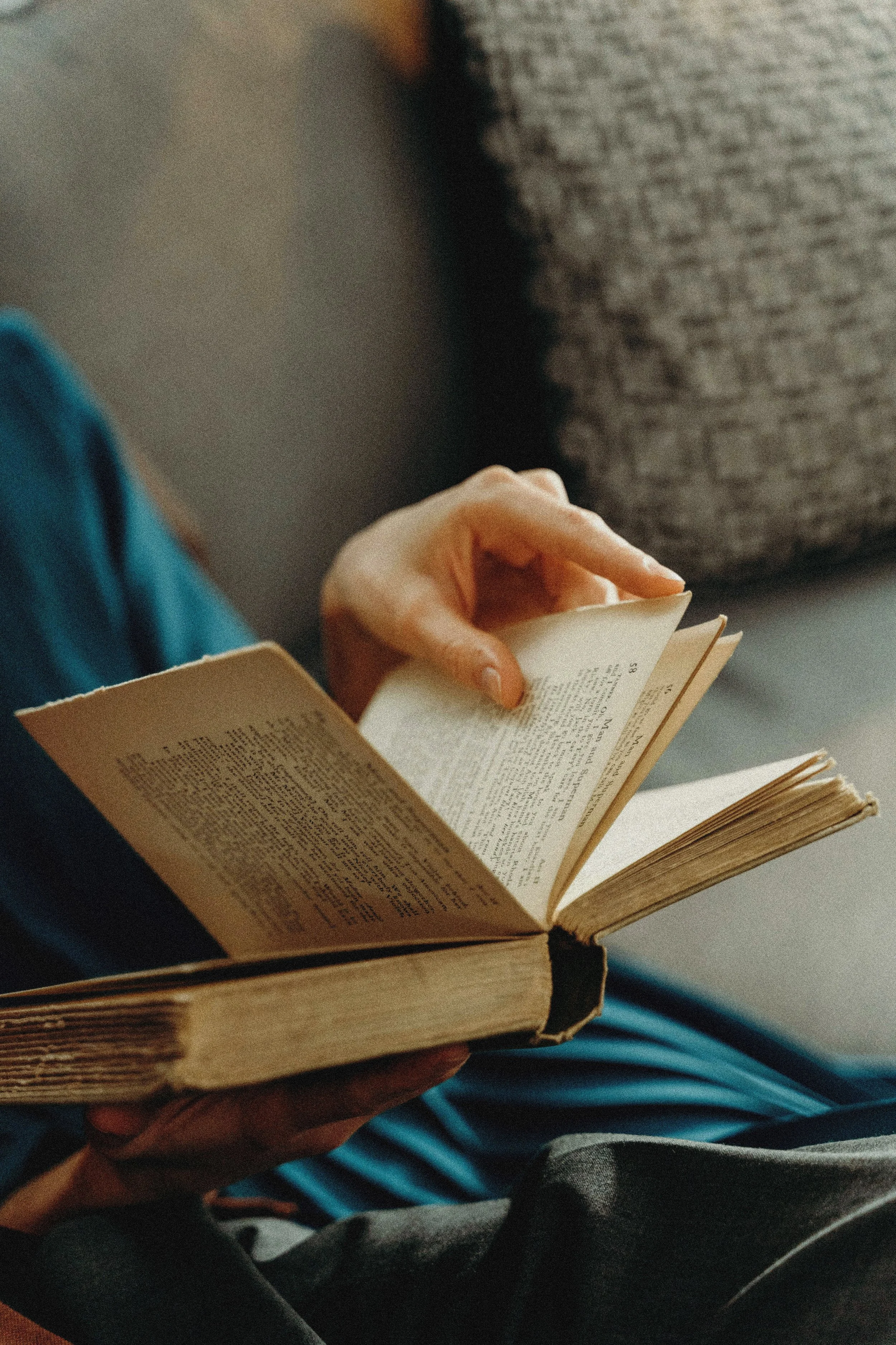 Close-up of a person reading a book, sitting on a couch with a pillow in the background.