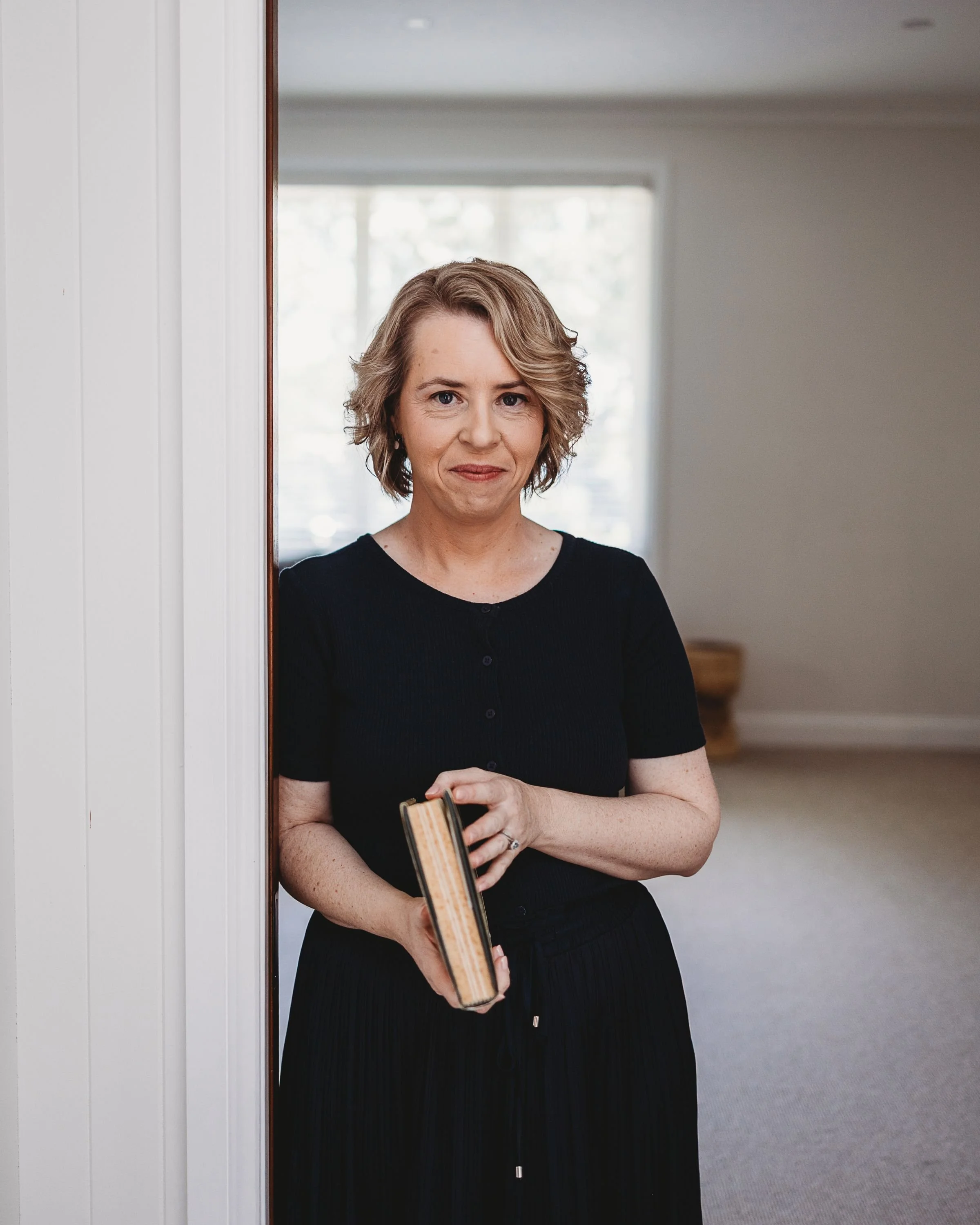 A woman with short, wavy blonde hair standing in a doorway, holding a closed book, smiling softly in a well-lit room with large windows.