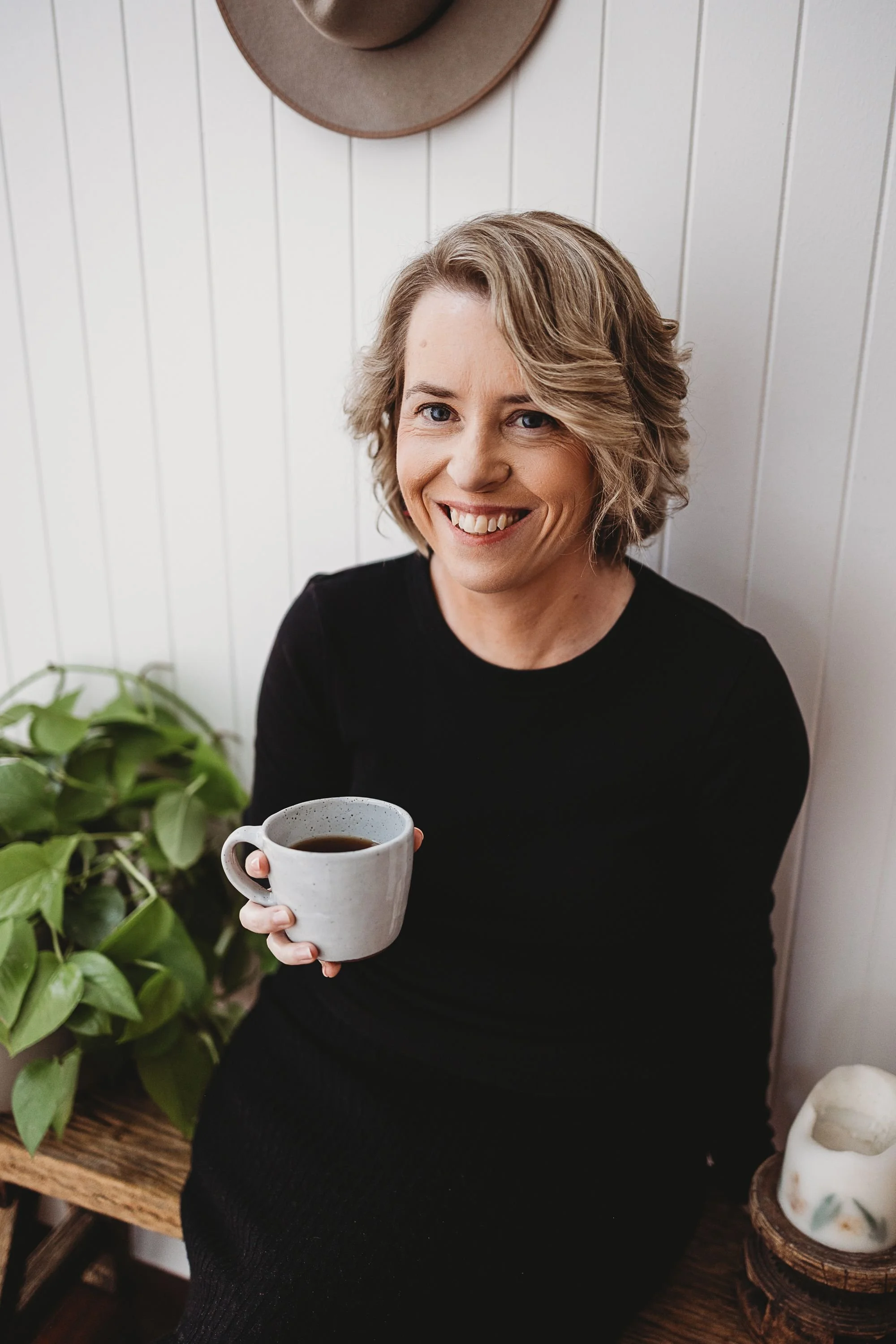 A woman with short, wavy, blonde hair, wearing a black top, smiling and holding a white mug filled with coffee, sitting near a houseplant on a wooden bench.