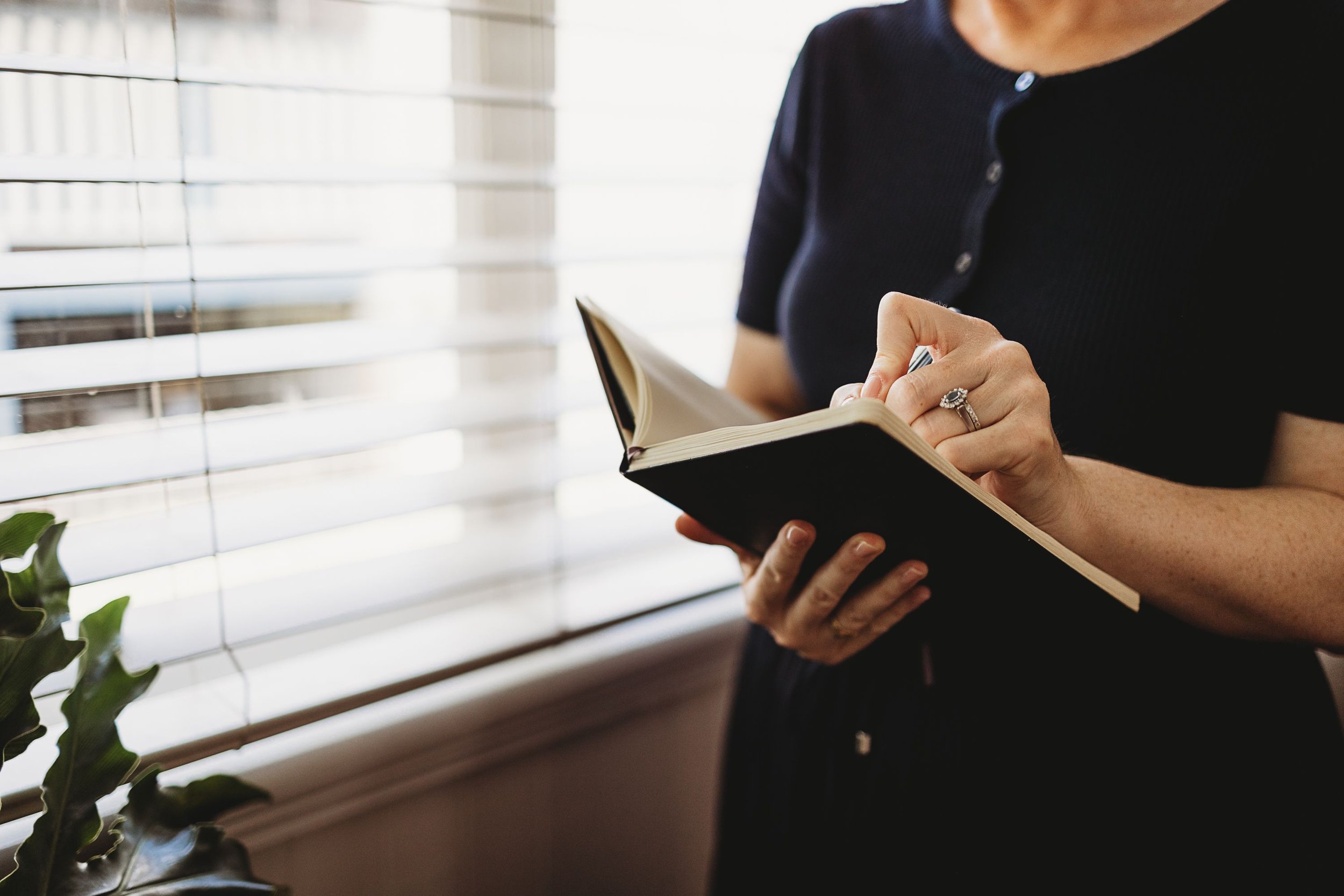 Person in black shirt holding a notebook near a window with blinds, reading or writing.