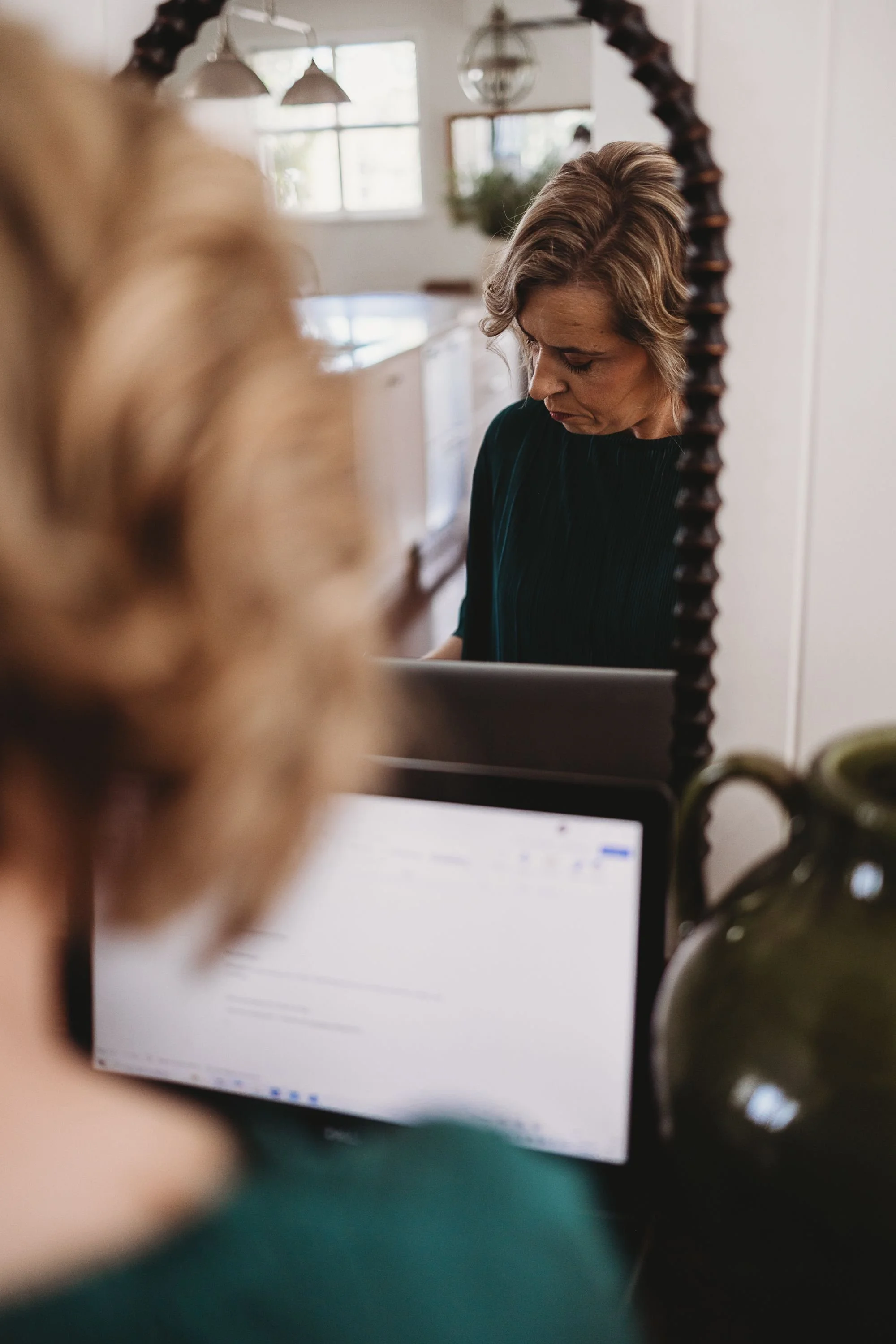 A woman with short, wavy hair looking down at a laptop, seen through a mirror, with another person's head and laptop in the foreground.