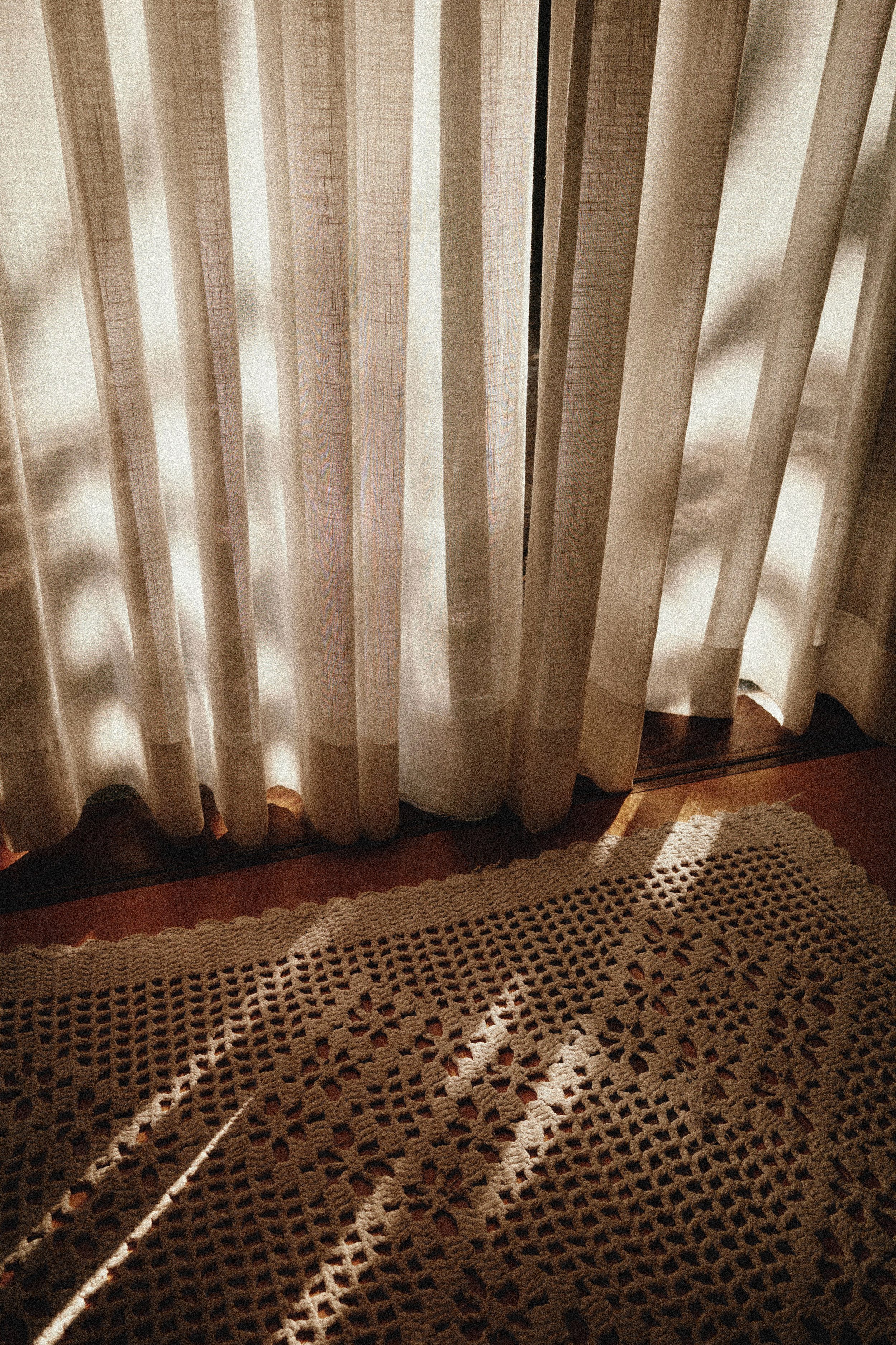 Curtains with sunlight casting shadows onto a crocheted rug on the floor.