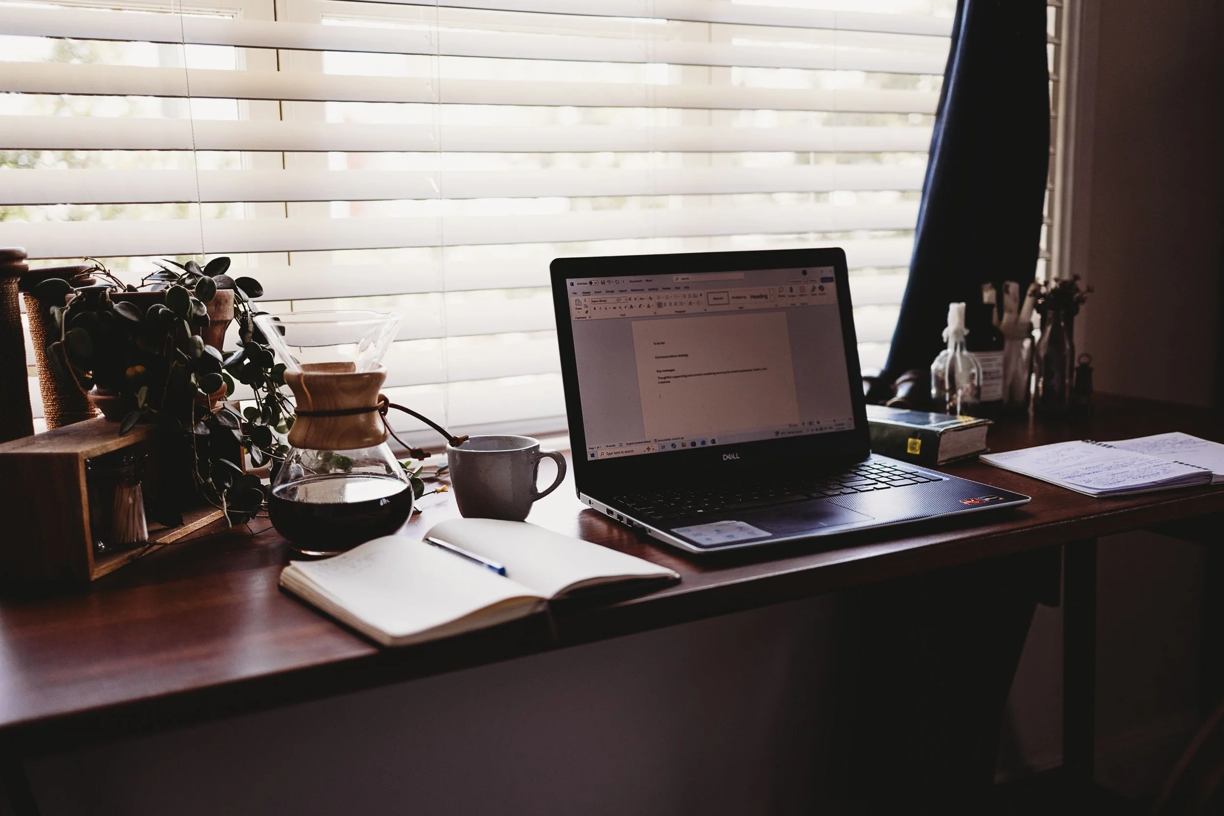 A desk with a laptop, an open notebook, a coffee pot, a mug, and various stationery items near window blinds letting in natural light.