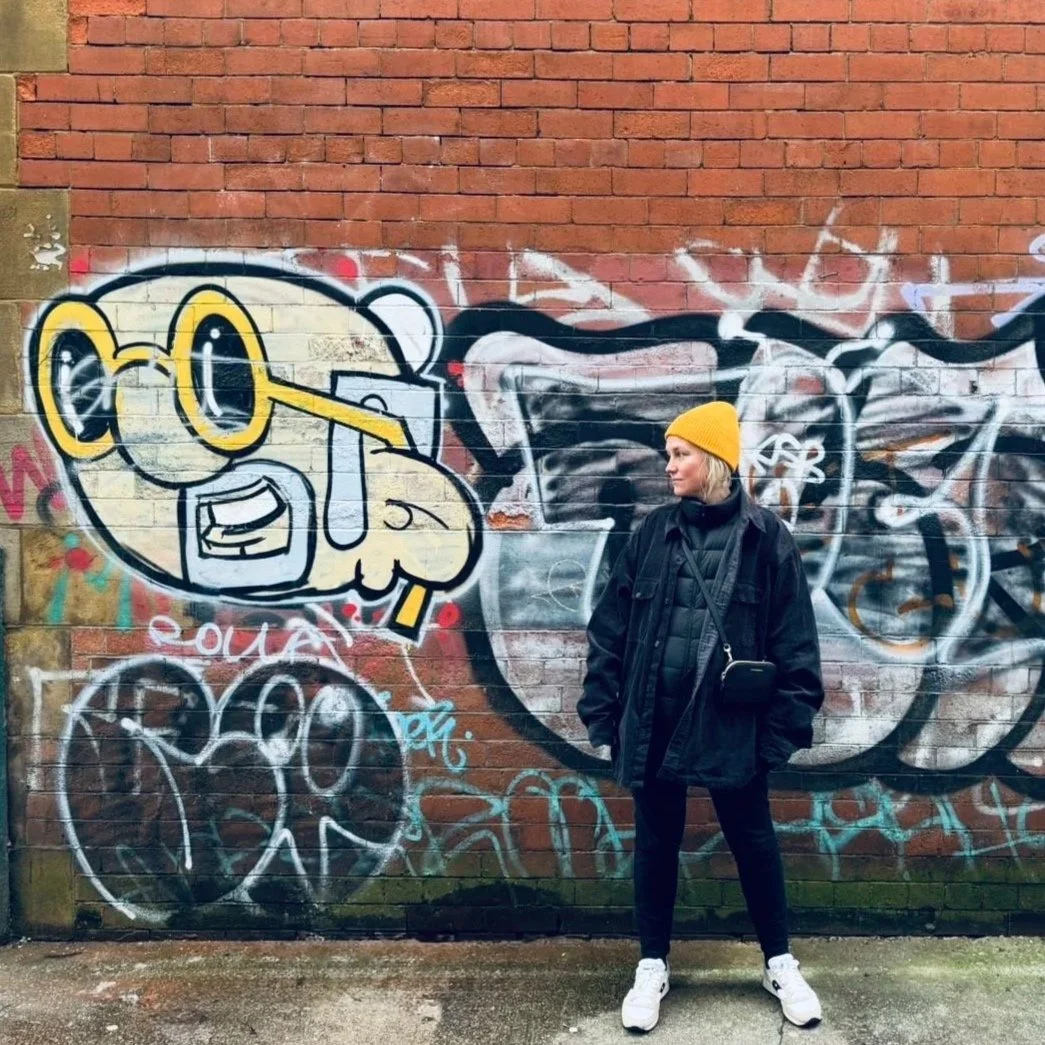 Woman wearing a yellow beanie, black jacket, and white sneakers standing in front of a graffiti mural on a brick wall.