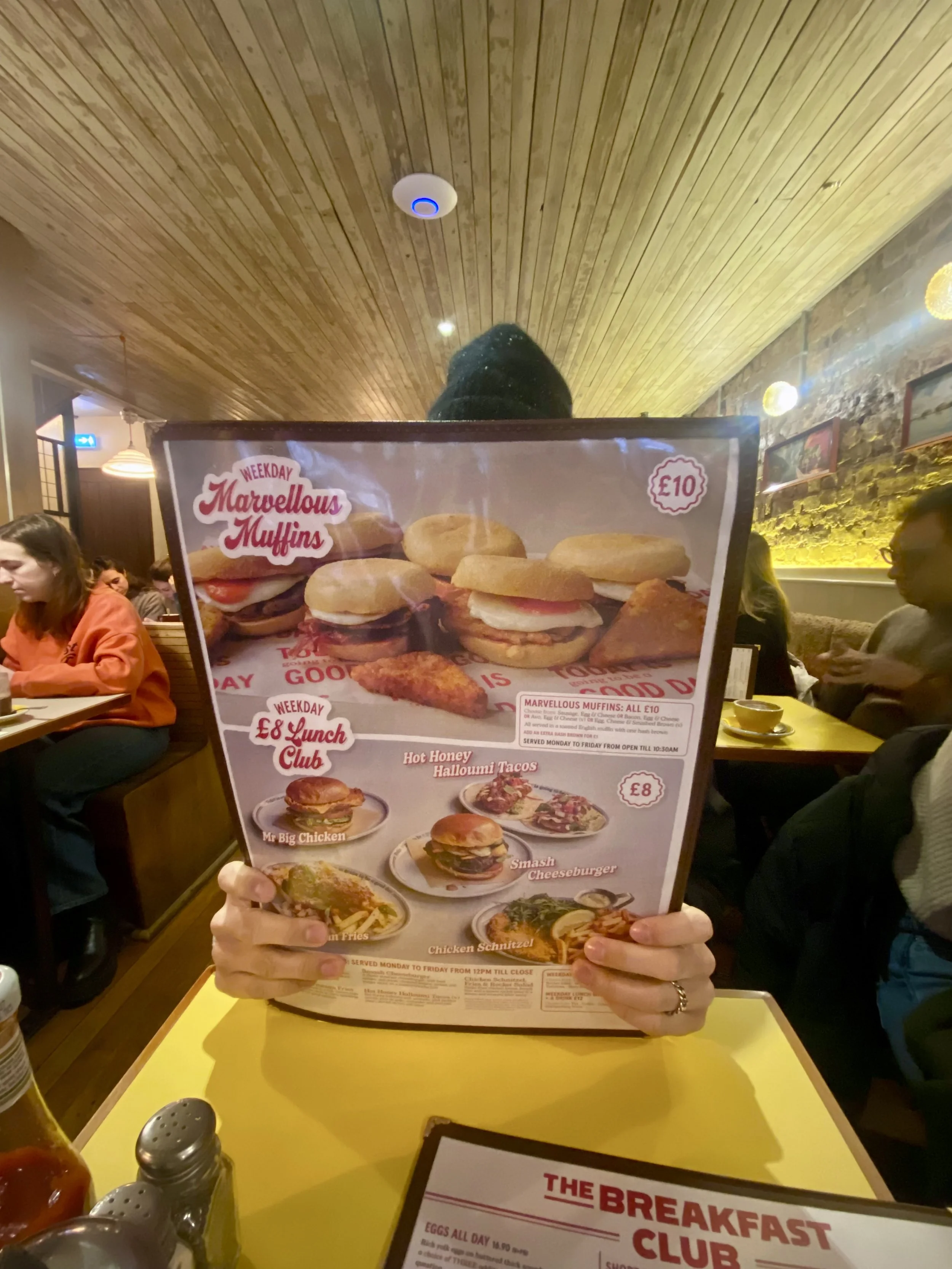 Person holding a restaurant menu featuring variety of muffins, burgers, fries, and other food items inside a cozy, warmly lit restaurant with wooden ceiling and brick walls.