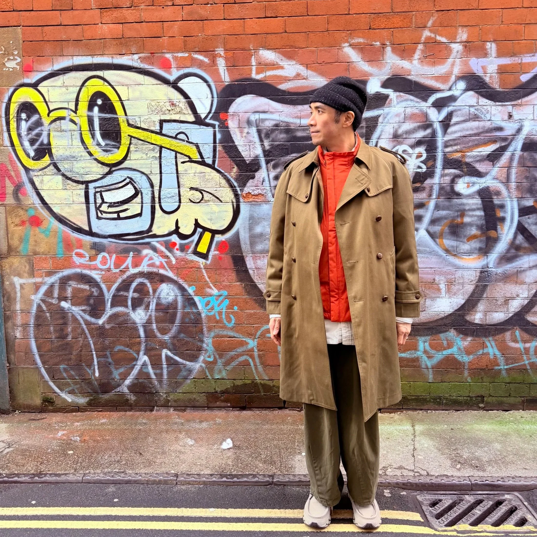 A man standing in front of a wall with colorful graffiti art, wearing a tan trench coat, orange jacket, black beanie, and beige sneakers.
