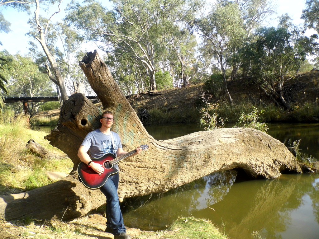 A young man standing in front of a large fallen tree by a river, playing an acoustic guitar, surrounded by trees and green foliage.