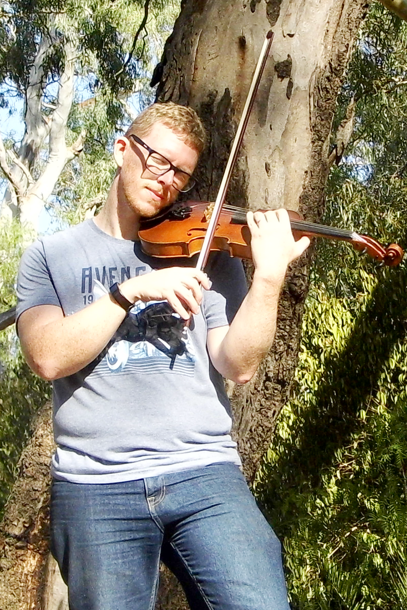A young man with glasses playing a violin outdoors in front of a tree with green leaves.