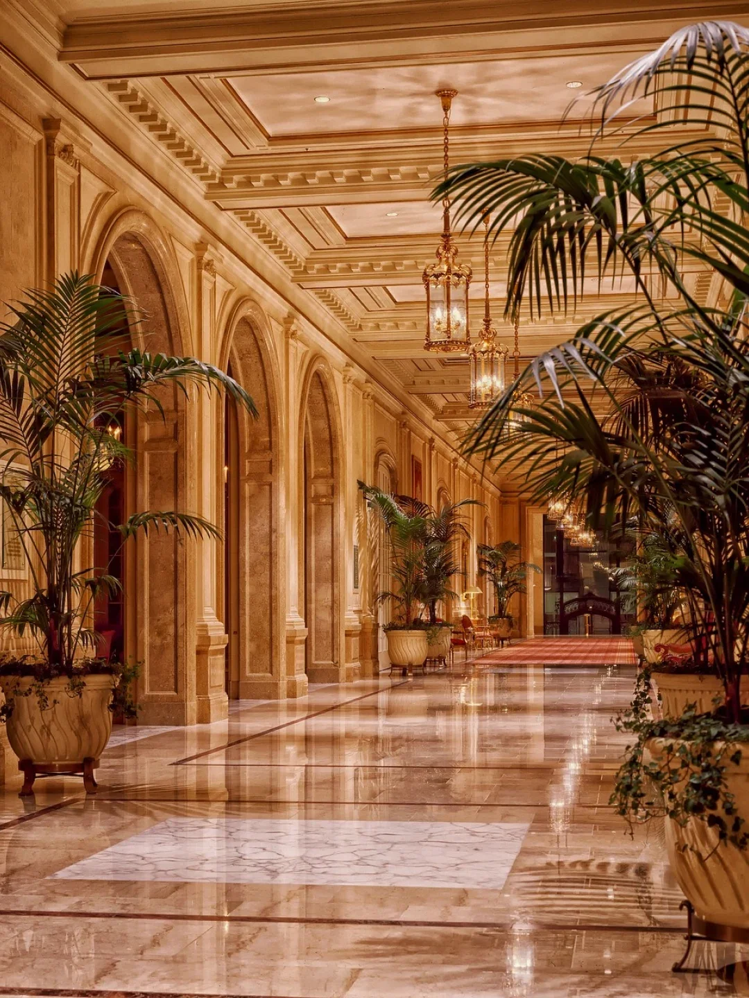 Luxurious hotel hallway with marble floors, ornate arches, gold hanging lanterns, and potted plants.