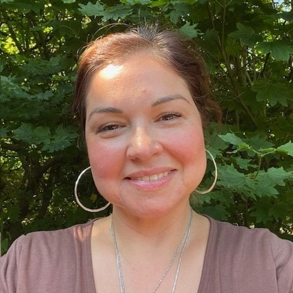 A smiling woman with hoop earrings and a necklace, standing outdoors in front of green leafy bushes.