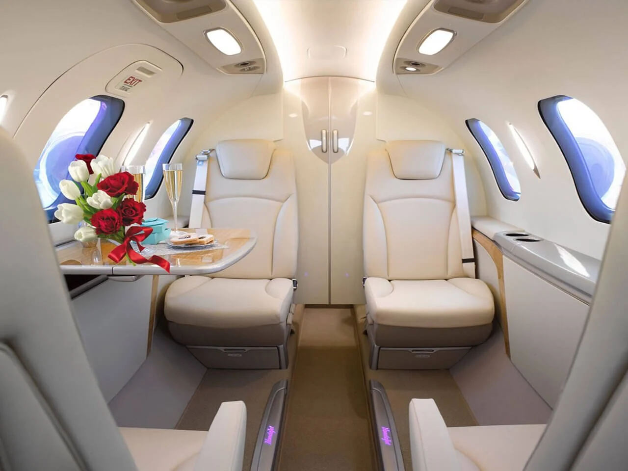 Interior of a luxury HondaJet with beige leather seats and a small table. On the table, there are red and white roses, two champagne glasses, and plates with snacks. The cabin is well-lit with large windows.